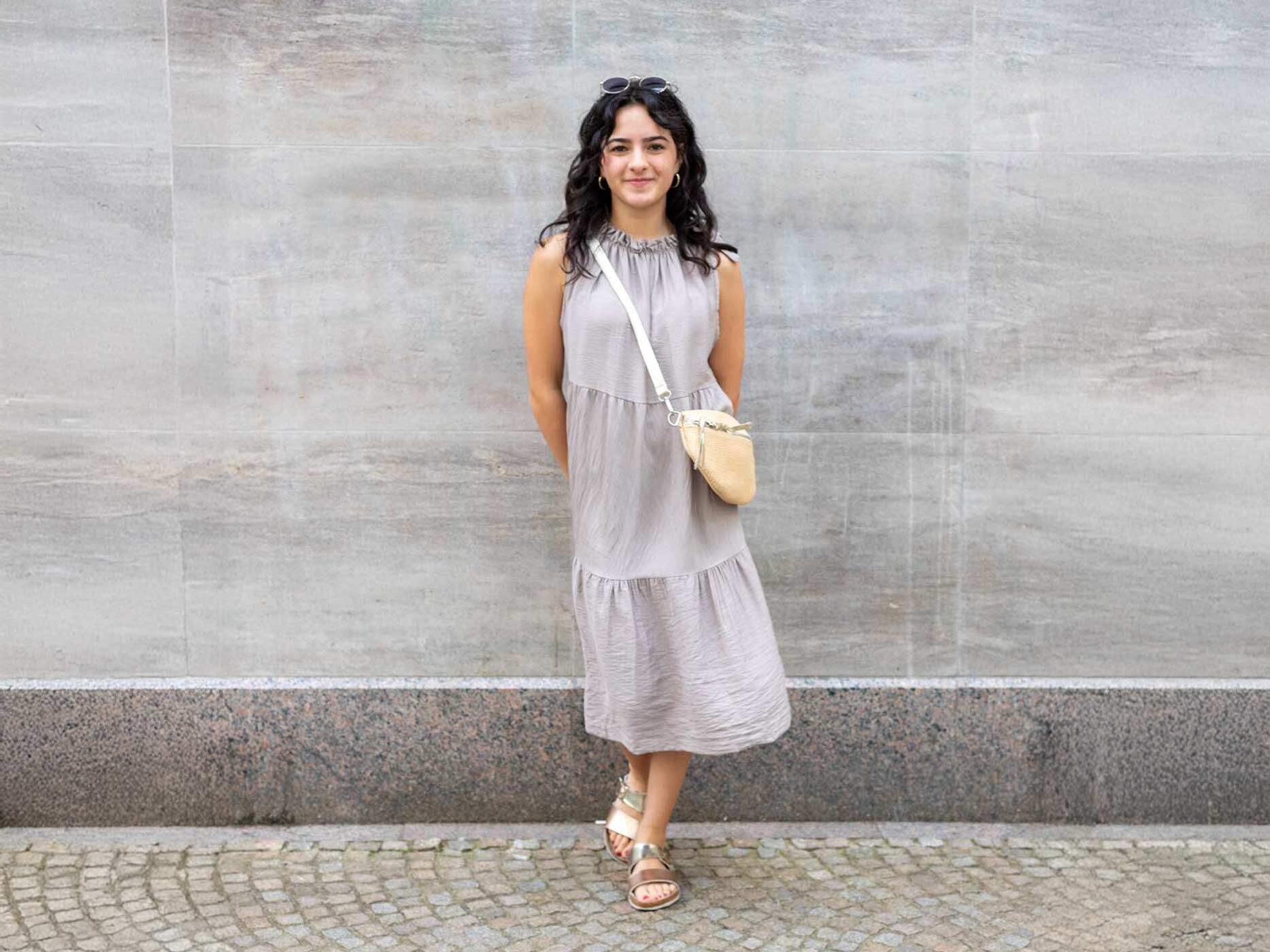 Eine Frau trägt ein greiges langes Sommerkleid, dazu eine graue Sonnenbrille, silberne Ohrringe und eine beige Handtasche und beige Sandalen.