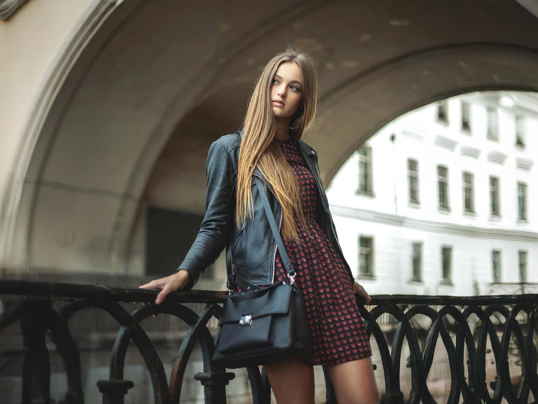 Eine Frau lehnt an einem Geländer, sie trägt ein kurzes schwarzes Kleid mit kleinen roten Herzen und dazu eine schwarze Lederjacke und eine schwarze Handtasche.