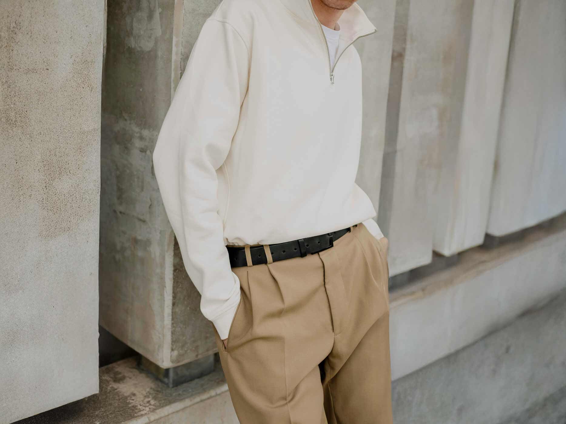 Ein Mann lehnt an einer Wand und er tr&auml;gt eine beige Hose und ein wei&szlig;es Sweatshirt mit einem Rei&szlig;verschluss