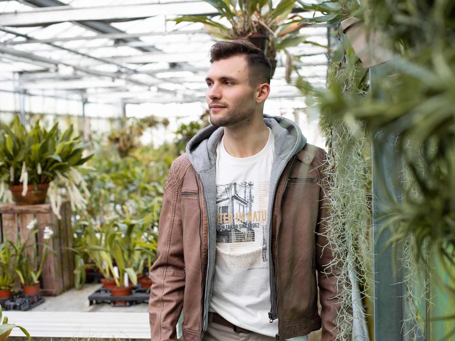 Ein Mann läuft durch ein Gewächshaus, er trägt ein weißes Oberteil mit Print und dazu eine graue Stoffjacke und darüber eine braune Lederjacke.
