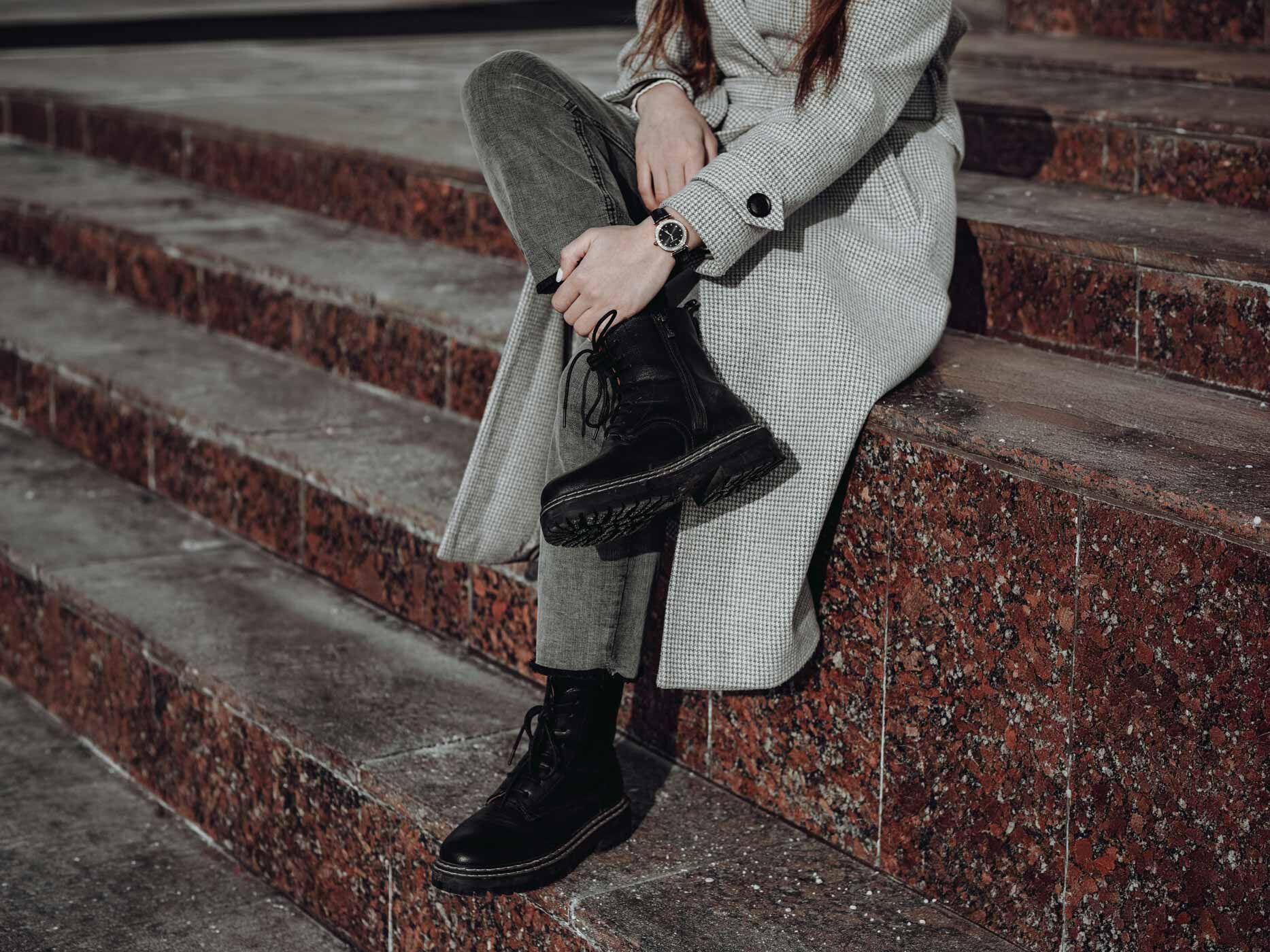 Eine Frau sitzt auf Treppenstufen, sie tr&auml;gt eine graue weitere Jeans wird mit robusten Schn&uuml;rboots und einem langen grauen Mantel.