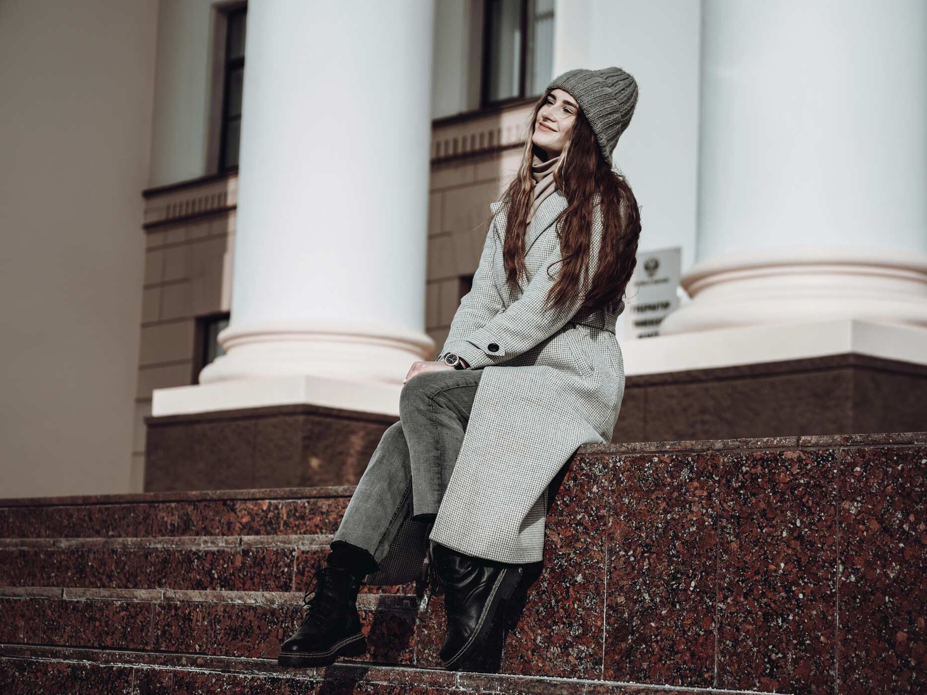 Eine Frau sitzt auf einer Treppe und guckt in den Himmel, sie trägt eine hellgraue Jeans, schwarze Boots, einen hellgrauen Mantel und eine dunkelgraue Mütze.