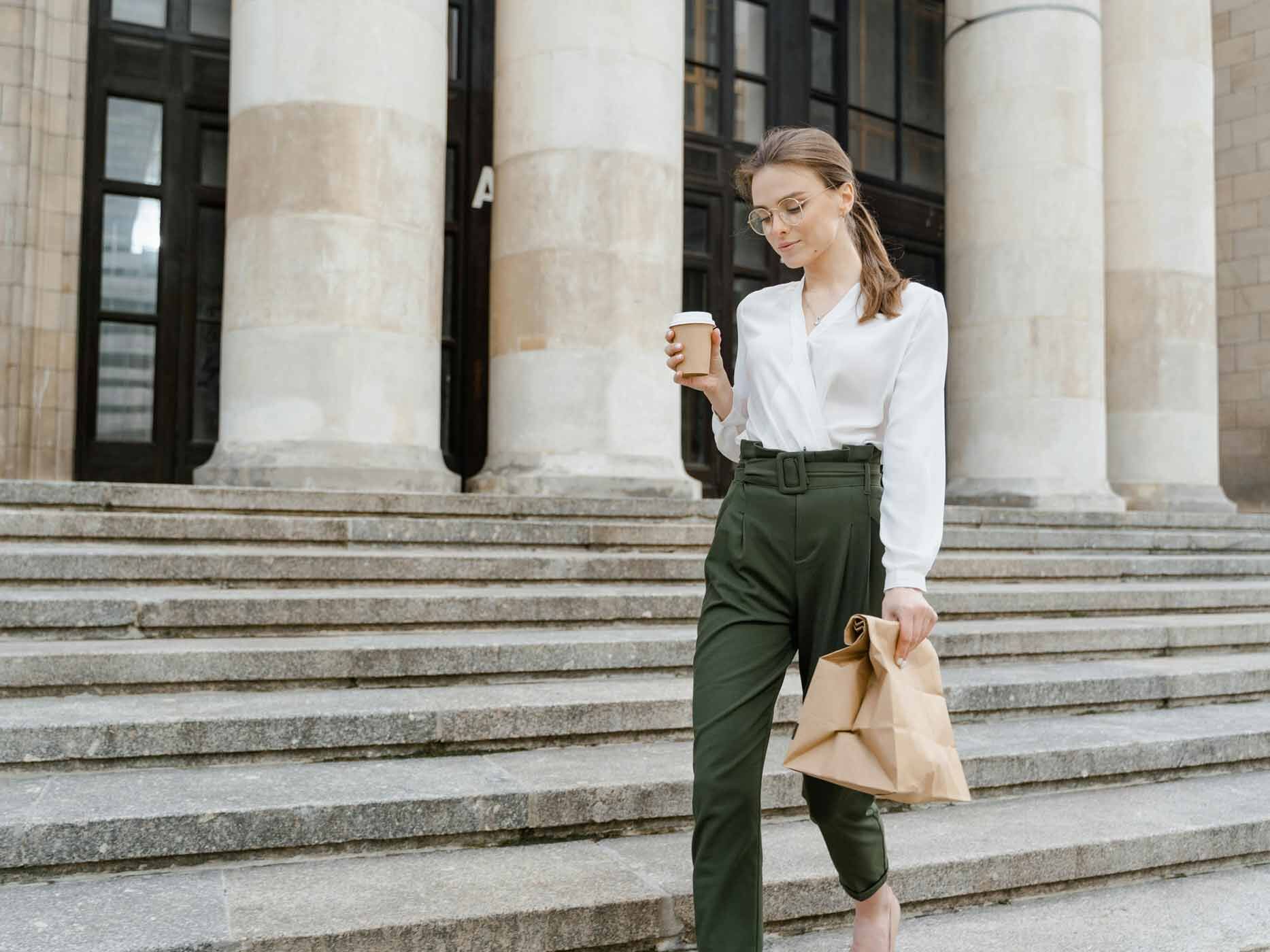 Eine Frau trägt eine grüne Stoffhose und eine weiße Bluse, sie hält einen Kaffee und eine braune Tüte in den Händen und geht eine Treppe entlang.