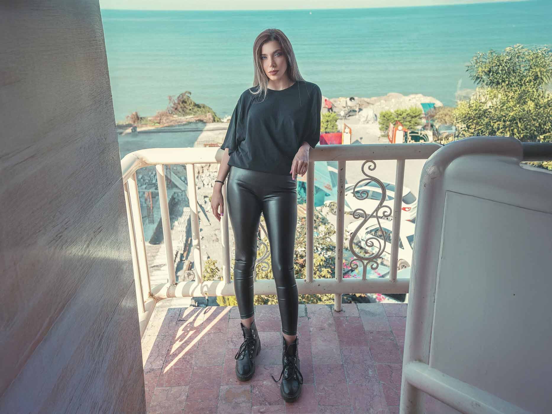 Eine Frau posiert auf einem Balkon und trägt eine enge, schwarze Lederjeans und ein schwarzes T-Shirt.
