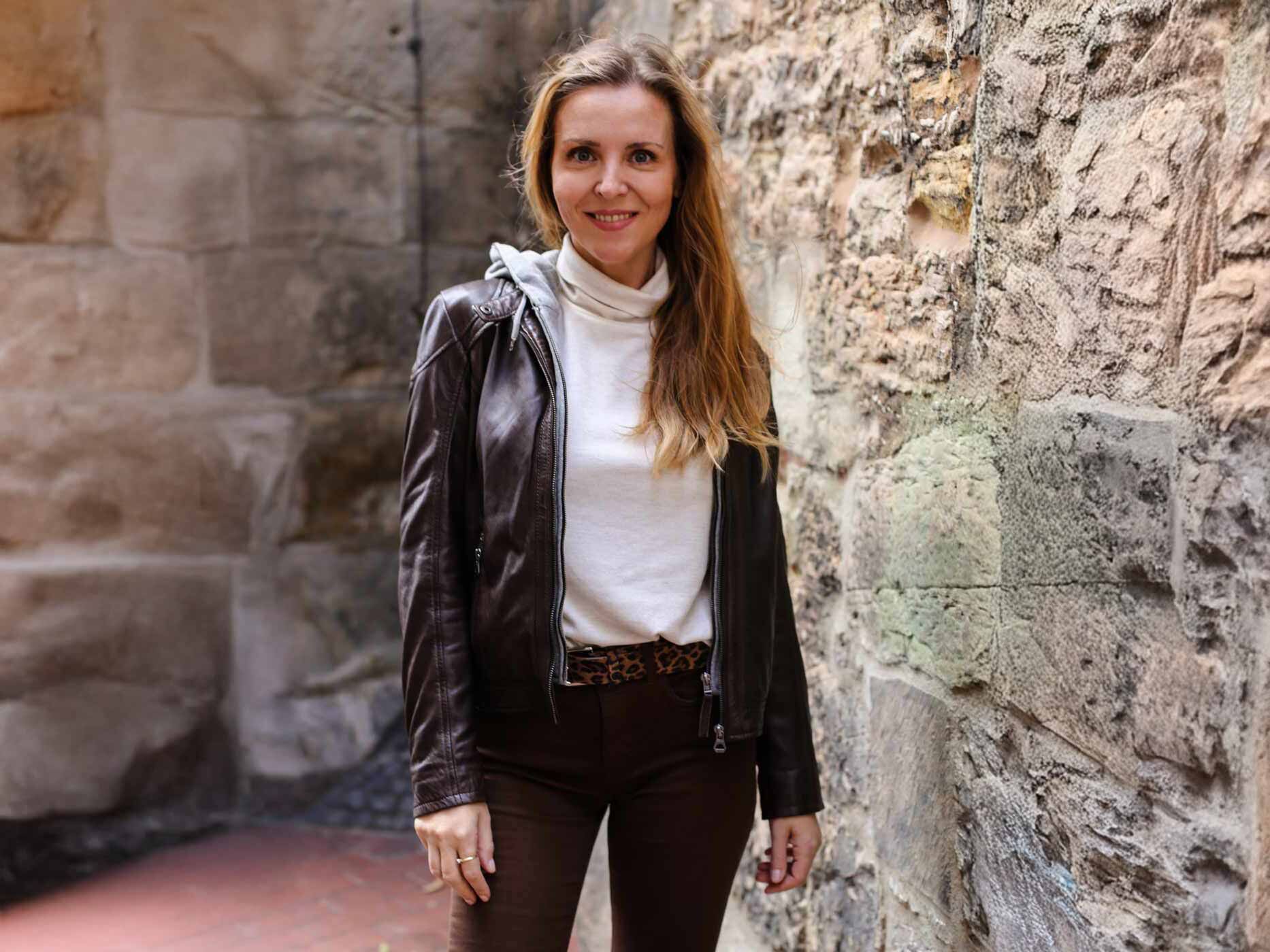 Eine Frau steht neben einer Steinwand, sie trägt eine braune Jeans, einen Leo-Gürtel, ein weißes Oberteil und dazu eine offene braune Lederjacke.