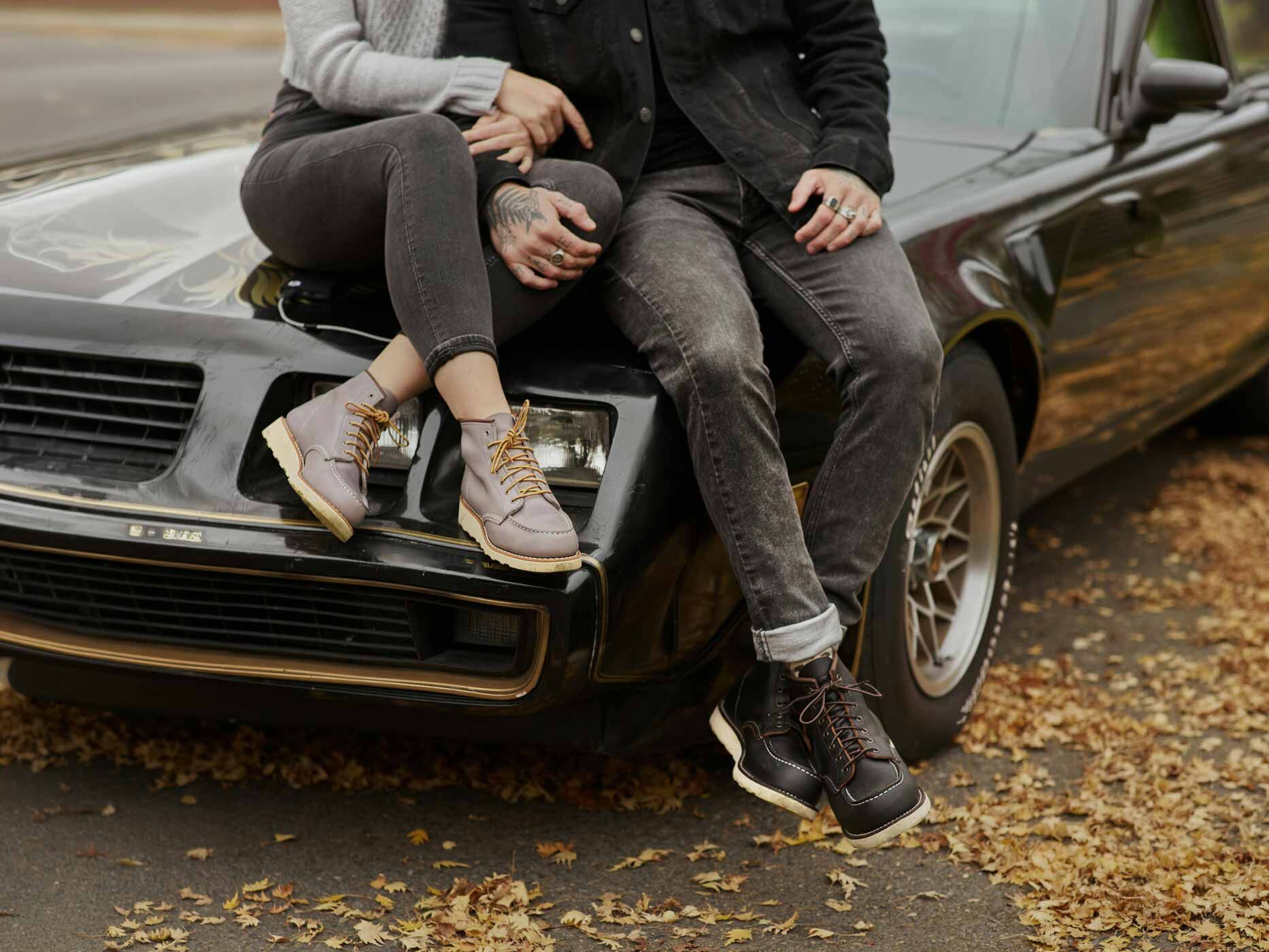 Eine Frau und ein Mann sitzen auf der Motorhaube eines Autos, doch man sieht nur ihre Beine, die Frau trägt eine schwarze Skinny Jeans mit braunen Boots.