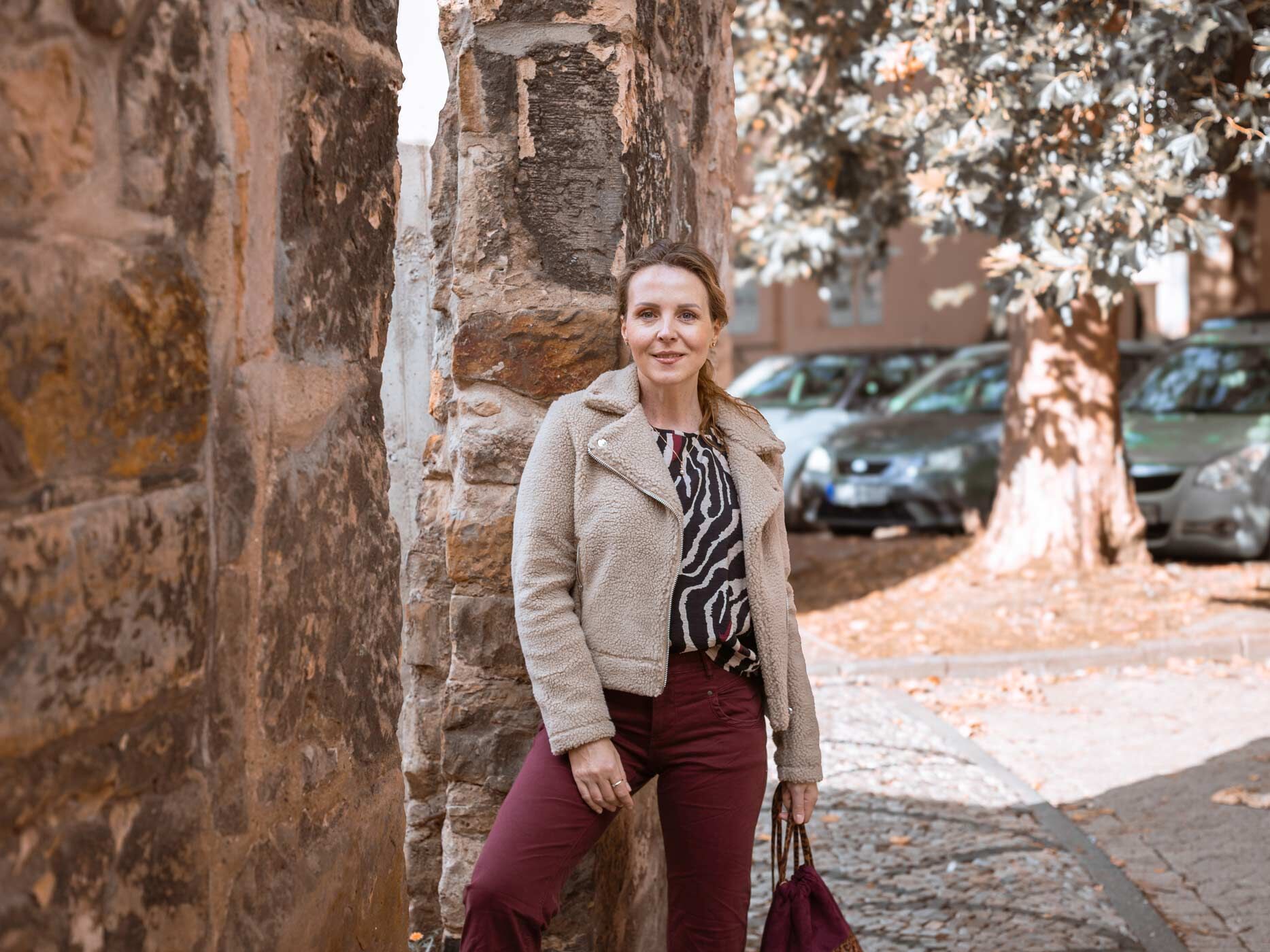 Eine Frau lehnt an einer Mauer, sie trägt eine dunkle, weinrote Hose, dazu ein beige-braunes Zebrastreifenoberteil und eine beige Teddyjacke.