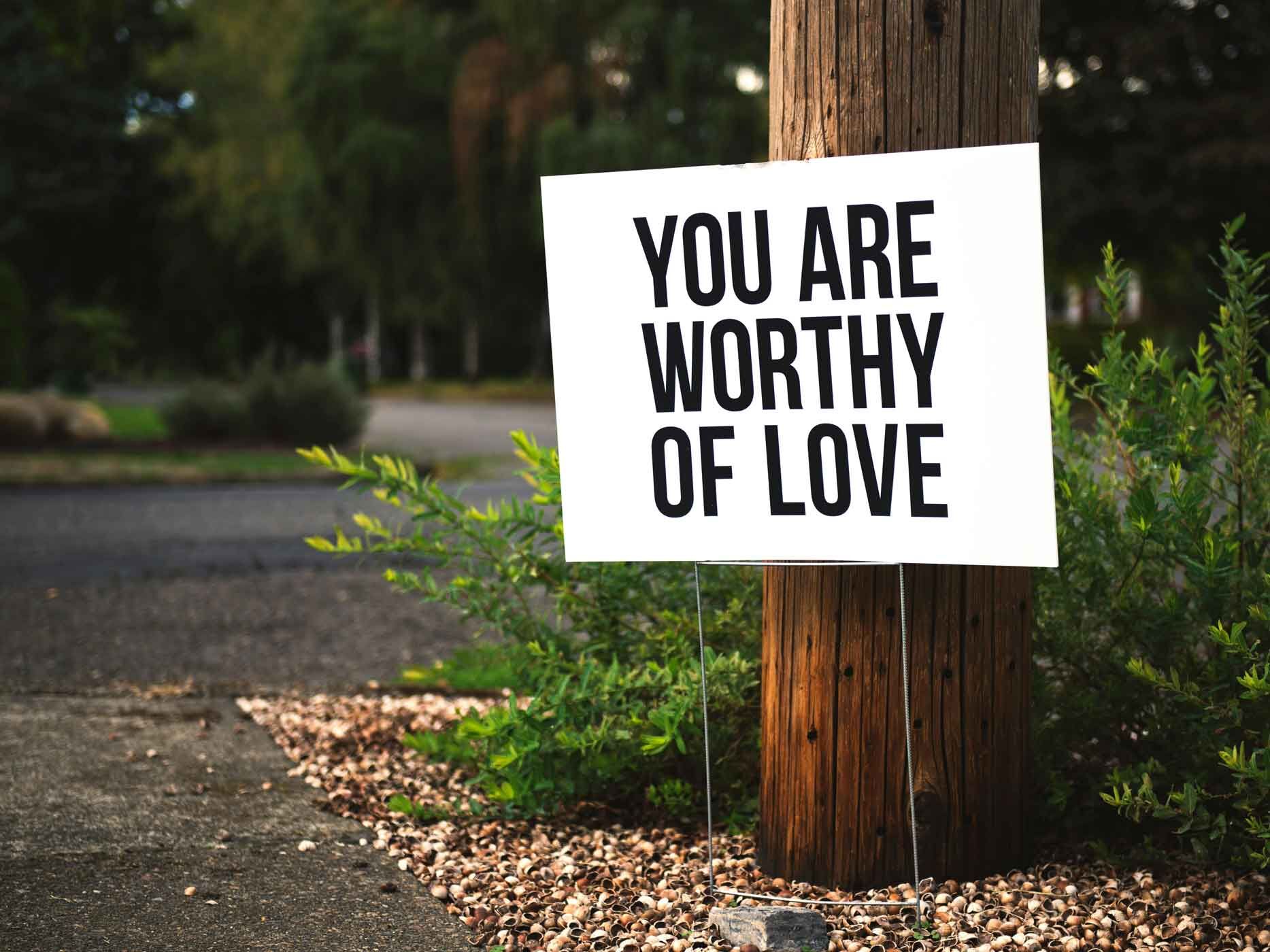 Vor einem Wald steht ein weißes Schild, auf dem "You are worthy of love" steht.