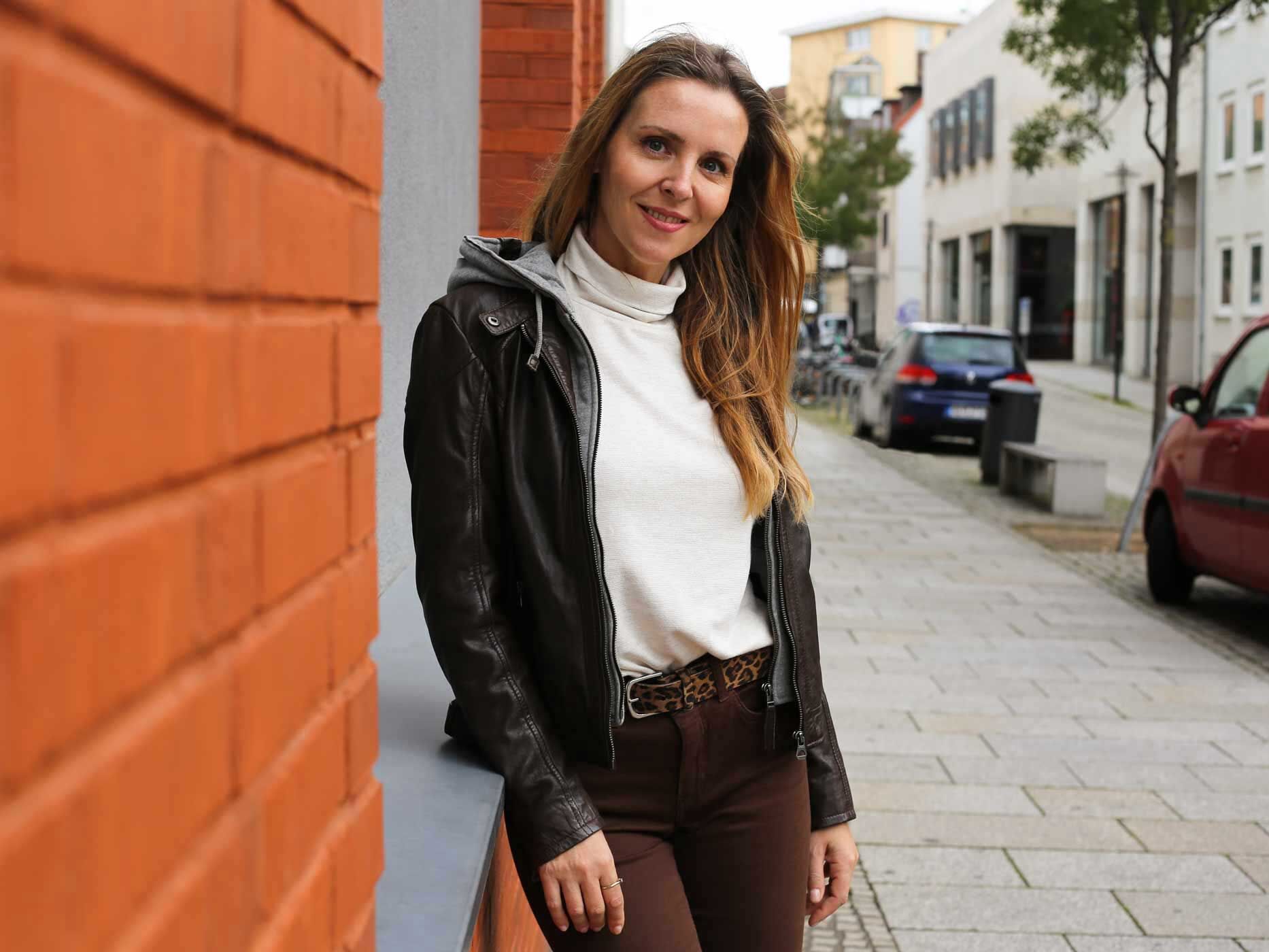 Eine Frau trägt eine braune Hose, einen weißen Pullover und eine schwarze Lederjacke. Dazu trägt sie einen Gürtel mit Leo-Print.