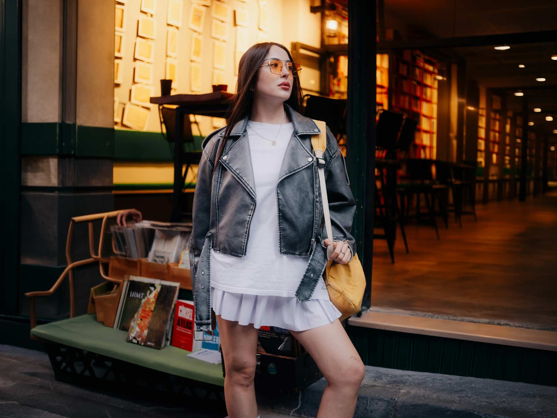 Eine Frau trägt einen kurzen weißen Rock, dazu ein weißes Oversize T-Shirt und darüber eine schwarze Lederjacke, sie trägt eine gelbe Handtasche und eine gelbe Sonnenbrille. Die Frau steht selbstbewusst vor einem stimmungsvoll beleuchteten Geschäft mit vielen Bücherregalen.