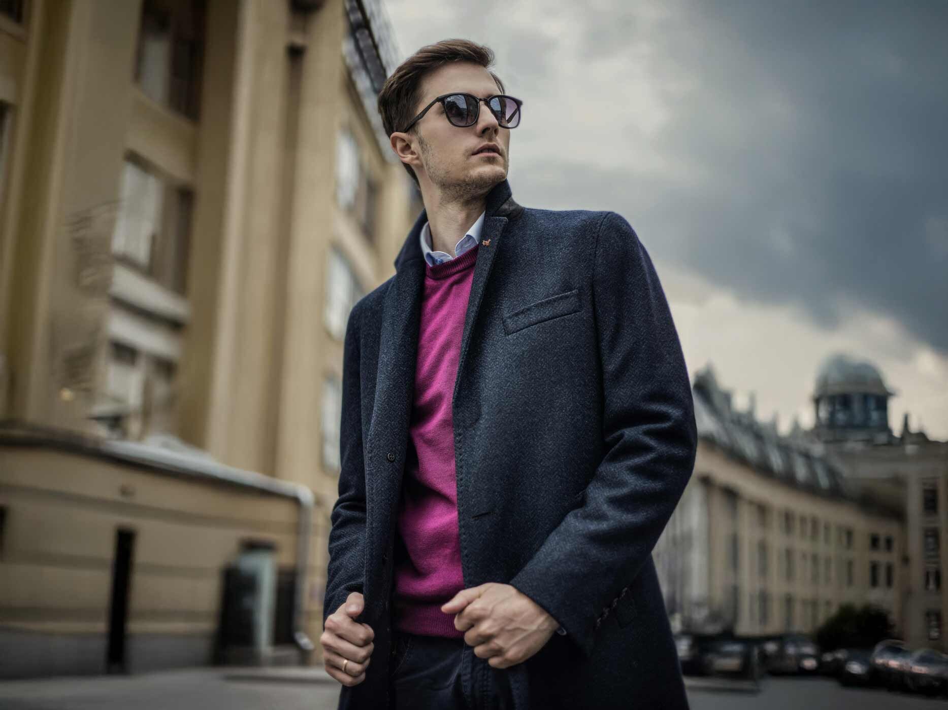 Ein Mann tr&auml;gt einen lilafarbenen Pullover, darunter ein wei&szlig;es Hemd und dar&uuml;ber einen dunkelgrauen Mantel und dazu eine Sonnenbrille.