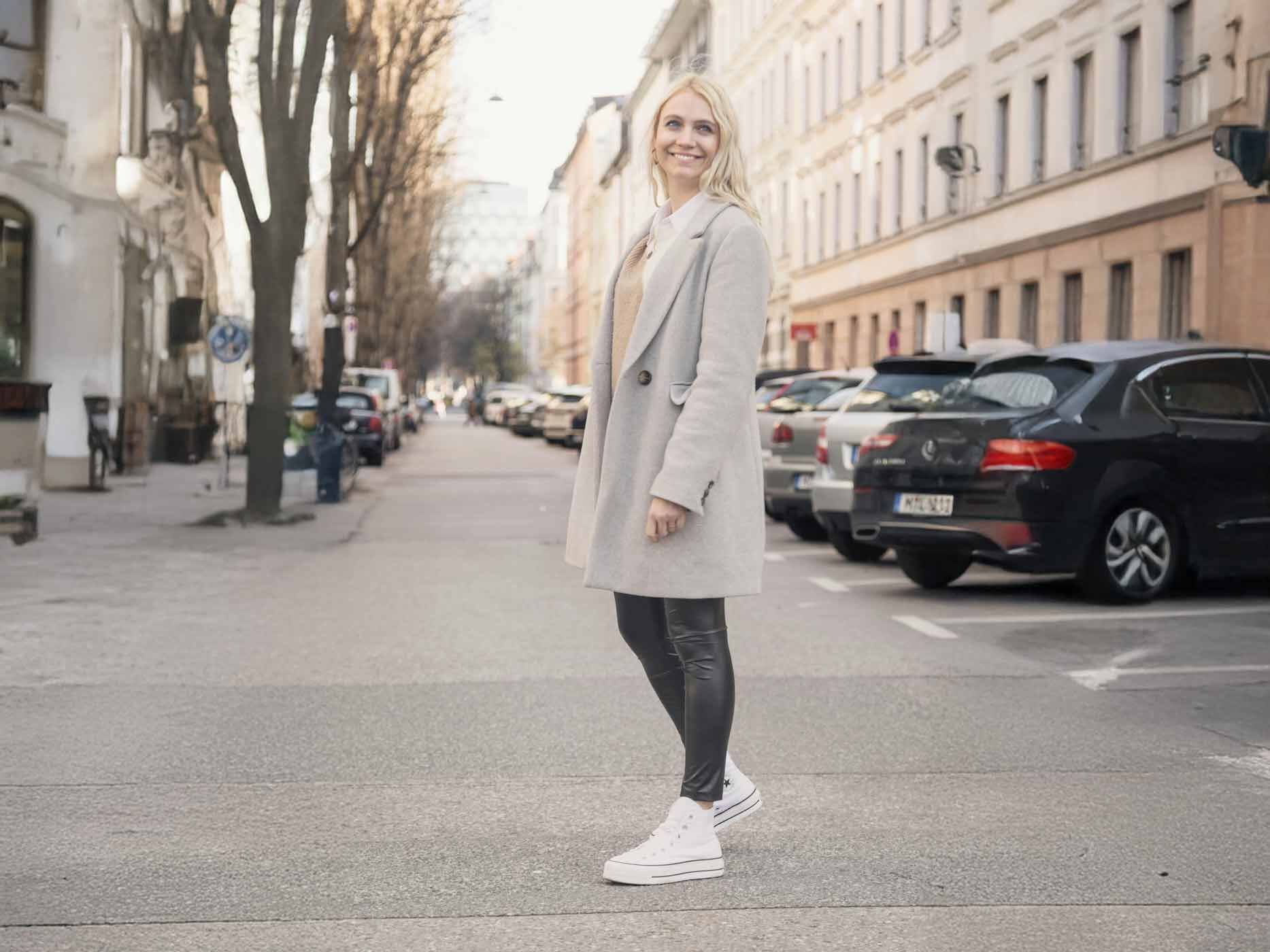 Eine Frau steht auf der Straße und trägt eine enge schwarze Lederhose und einen langen, grauen Mantel.