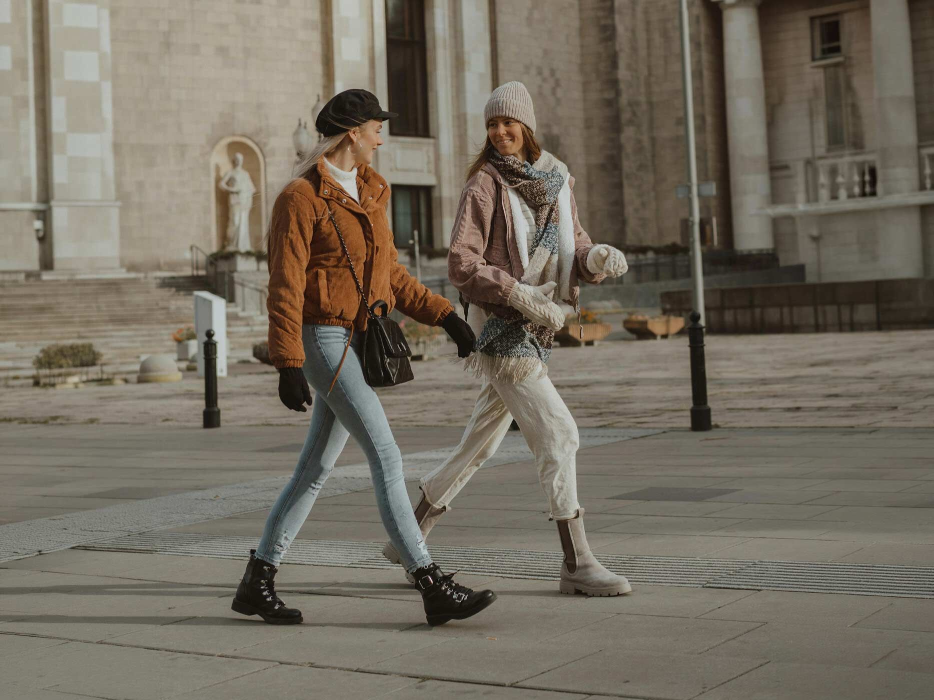 Zwei Frauen spazieren durch die Stad. Links ein lässiger Look mit brauner Steppjacke, heller Jeans und schwarzen Boots, kombiniert mit Cap und Umhängetasch, rechts ein softer, heller Style mit beiger Hose, Jacke, Schal und Strickmütze sowie robusten Boots in Naturtönen.