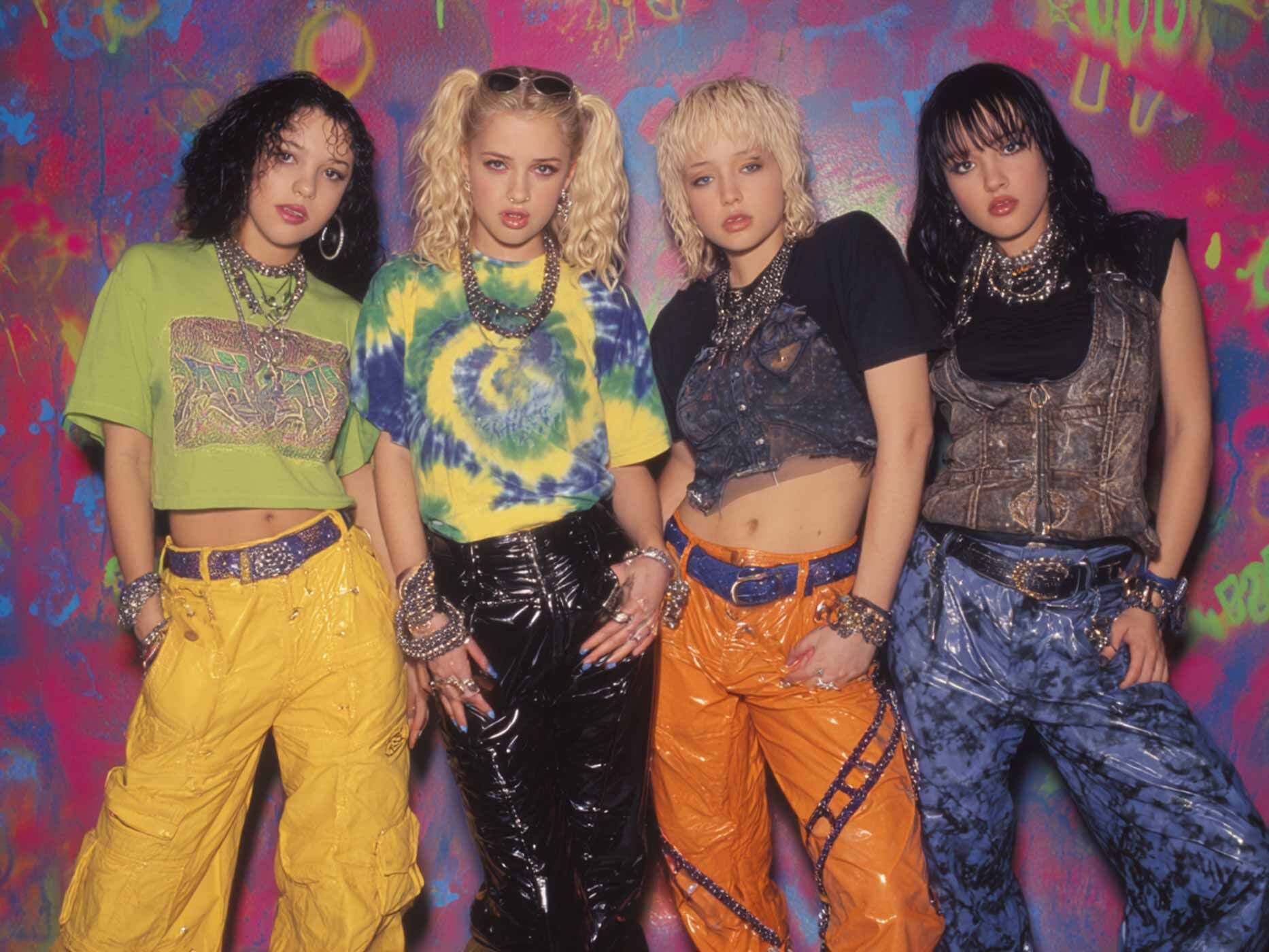 Vier junge Frauen stehen nebeneinander vor einer bunten, grafisch bemalten Wand. Sie tragen auff&auml;llige 90er Jahre Outfits mit bauchfreien Tops, weiten Hosen aus gl&auml;nzendem Material und kr&auml;ftigen Farben wie Gelb, Orange, Schwarz und Blau. Gro&szlig;e Ketten, Armreifen und G&uuml;rtel mit Nieten pr&auml;gen den Look. Die Frisuren reichen von Locken &uuml;ber Z&ouml;pfe bis zu kurzen, fransigen Schnitten. Alle blicken selbstbewusst in die Kamera.