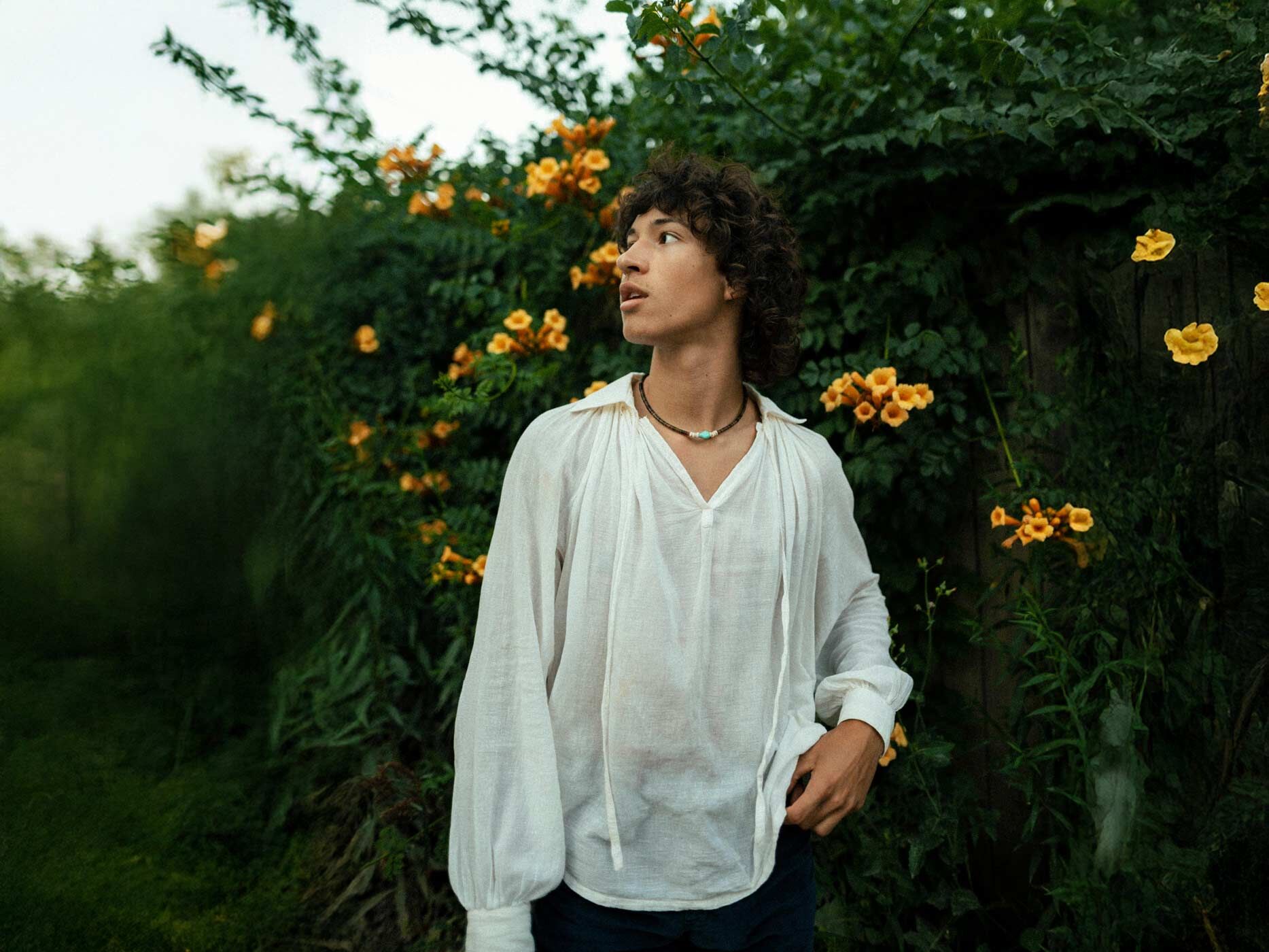 Ein junger Mann ein weißes, locker geschnittenes Hemd mit einem weiten Ausschnitt und posiert vor einer Hecke.