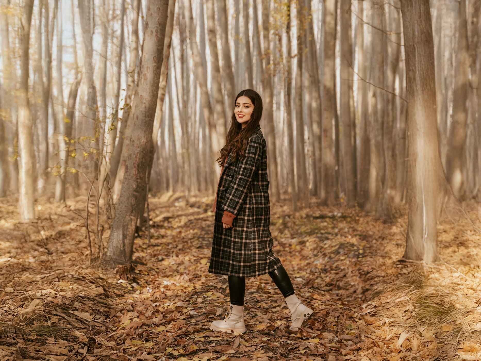 Eine Frau geht im Herbstwald spazieren und trägt eine enge schwarze Lederhose, hellbraune Stiefel und einen schwarz-braun karierten Mantel.