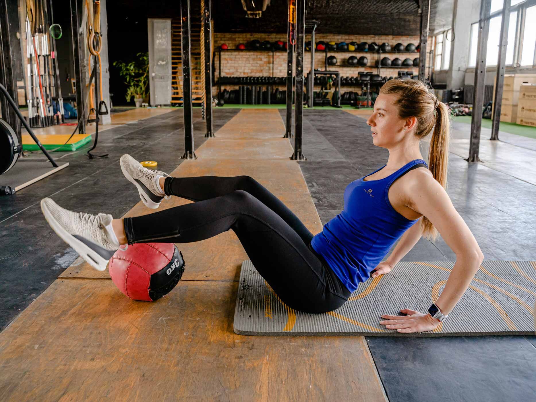 Eine Frau sitzt in Sportklamotten auf einer Sportmatte und hat ihre Beine auf einen großen Sportball gelegt.