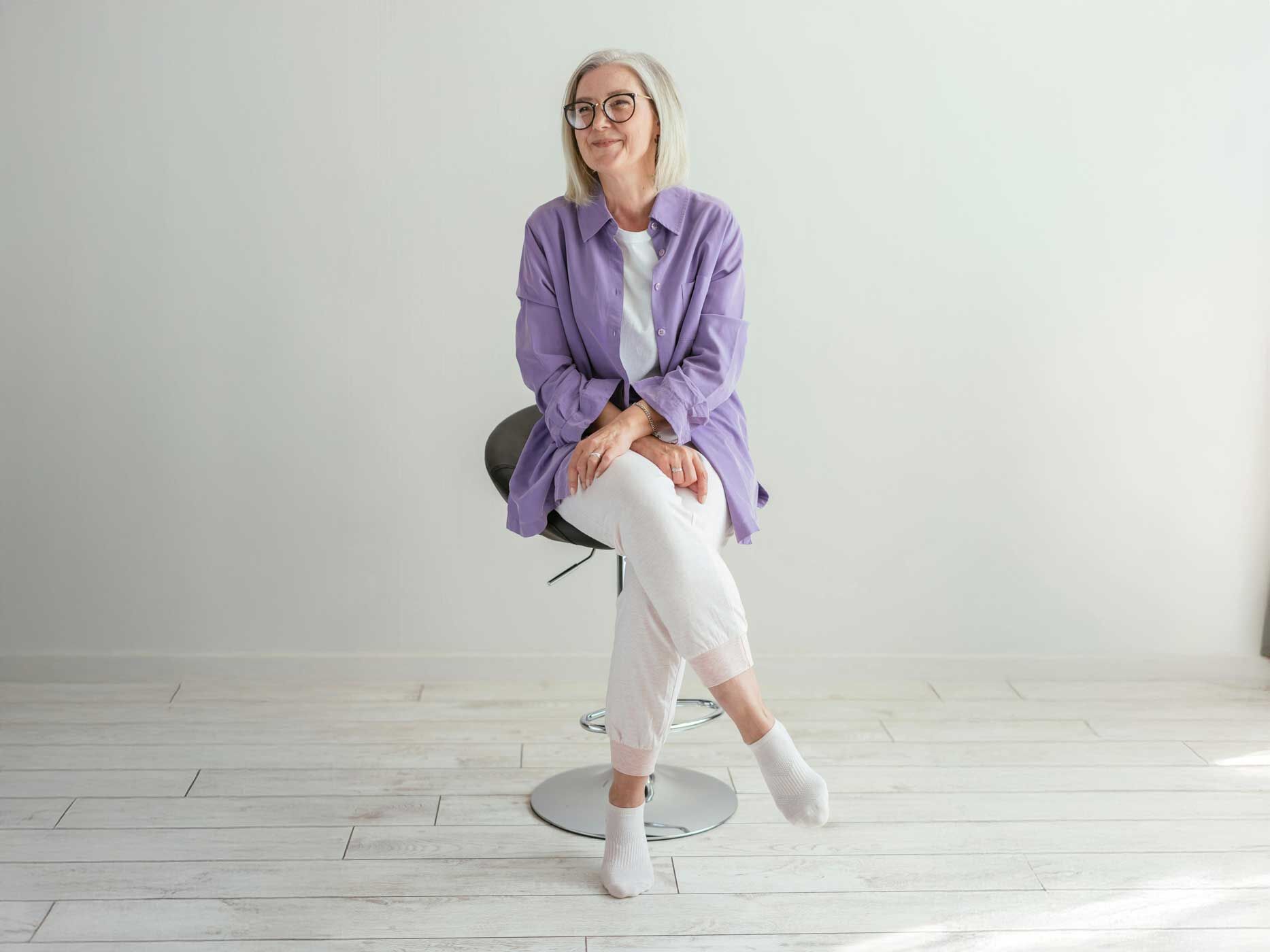 Eine Frau mit schulterlangen, grauen offenen Haaren sitzt auf einem Hocker, sie tr&auml;gt eine wei&szlig;e Hose, wei&szlig;e Socken und ein wei&szlig;es T-Shirt, dar&uuml;ber tr&auml;gt sie ein offenes lila Hemd und eine schwarze Brille.