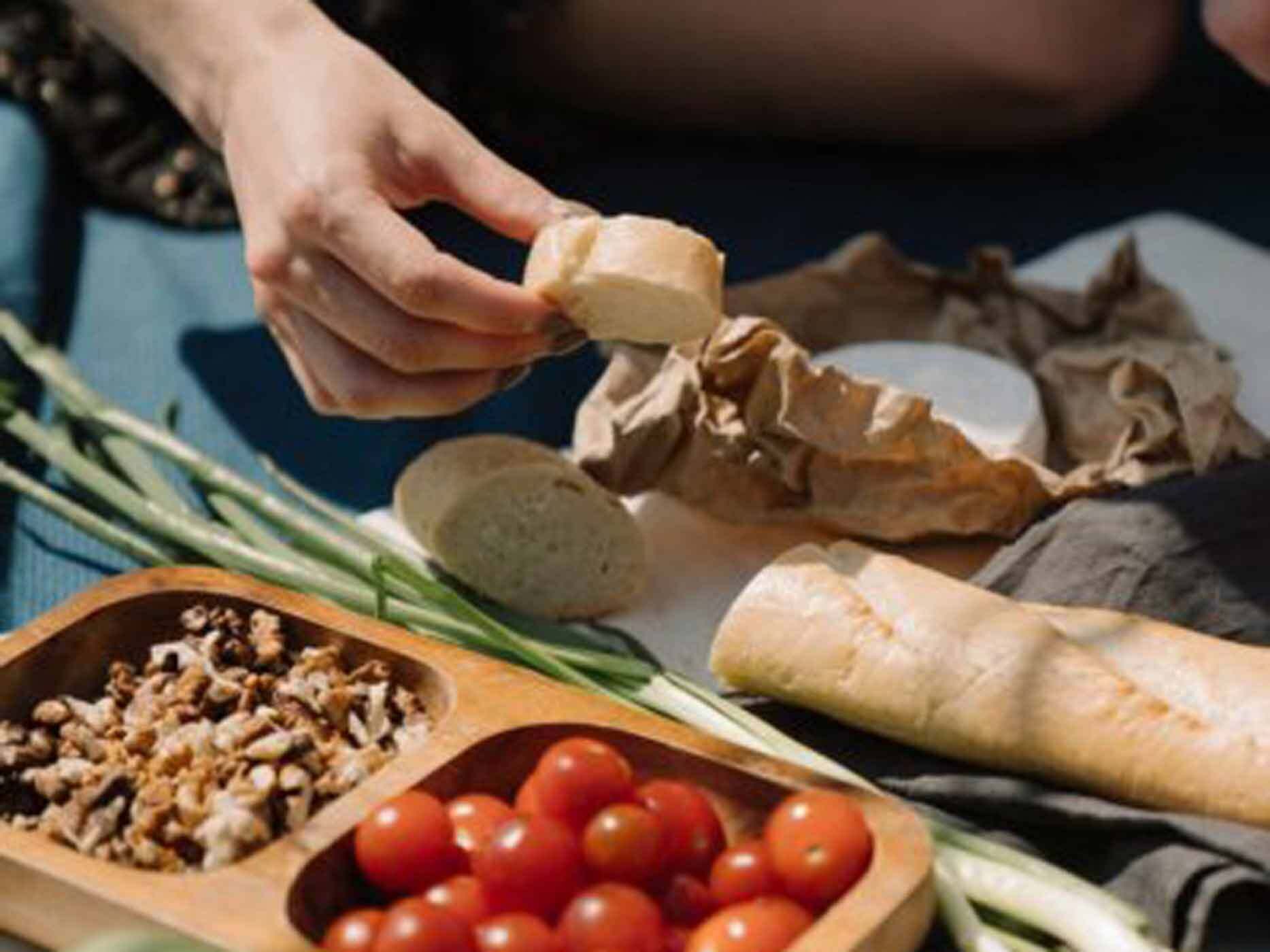Eine Hand h&auml;lt ein geschnittenes St&uuml;ck Baguette in der Hand, daneben steht ein runder K&auml;se, eine Dose mit N&uuml;ssen und Tomaten.