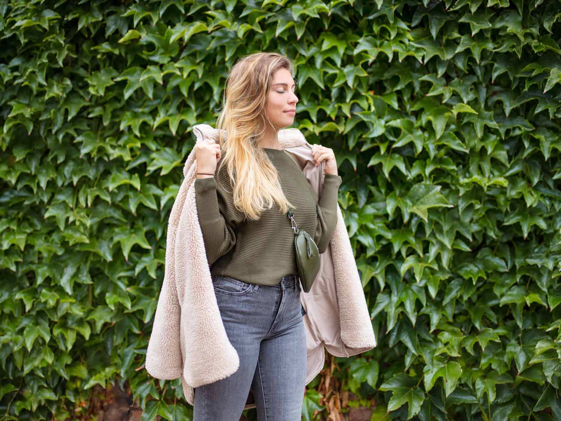 Eine Frau steht vor einer Hecke, sie trägt eine hellgraue Hose, einen dunkelgrünen Pullover und eine gleichfarbige Bauchtasche und dazu eine beige Teddyjacke.