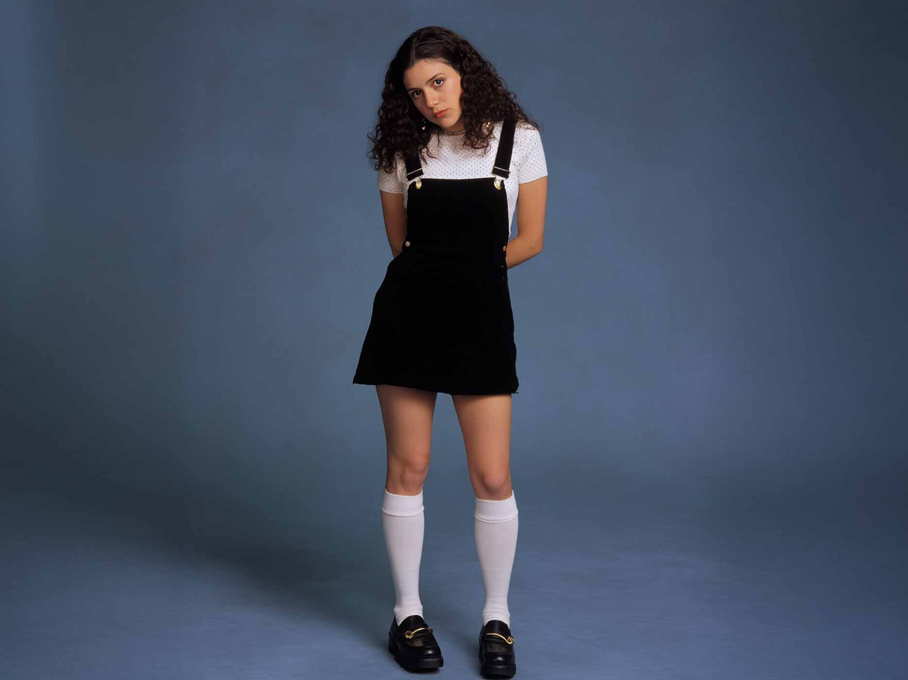 Eine junge Erwachsene posiert für die Kamera, sie trägt schwarze Schuhe, weiße lange Kniestrümpfe, ein weißes T-Shirt und darüber ein schwarzes Slip Dress.