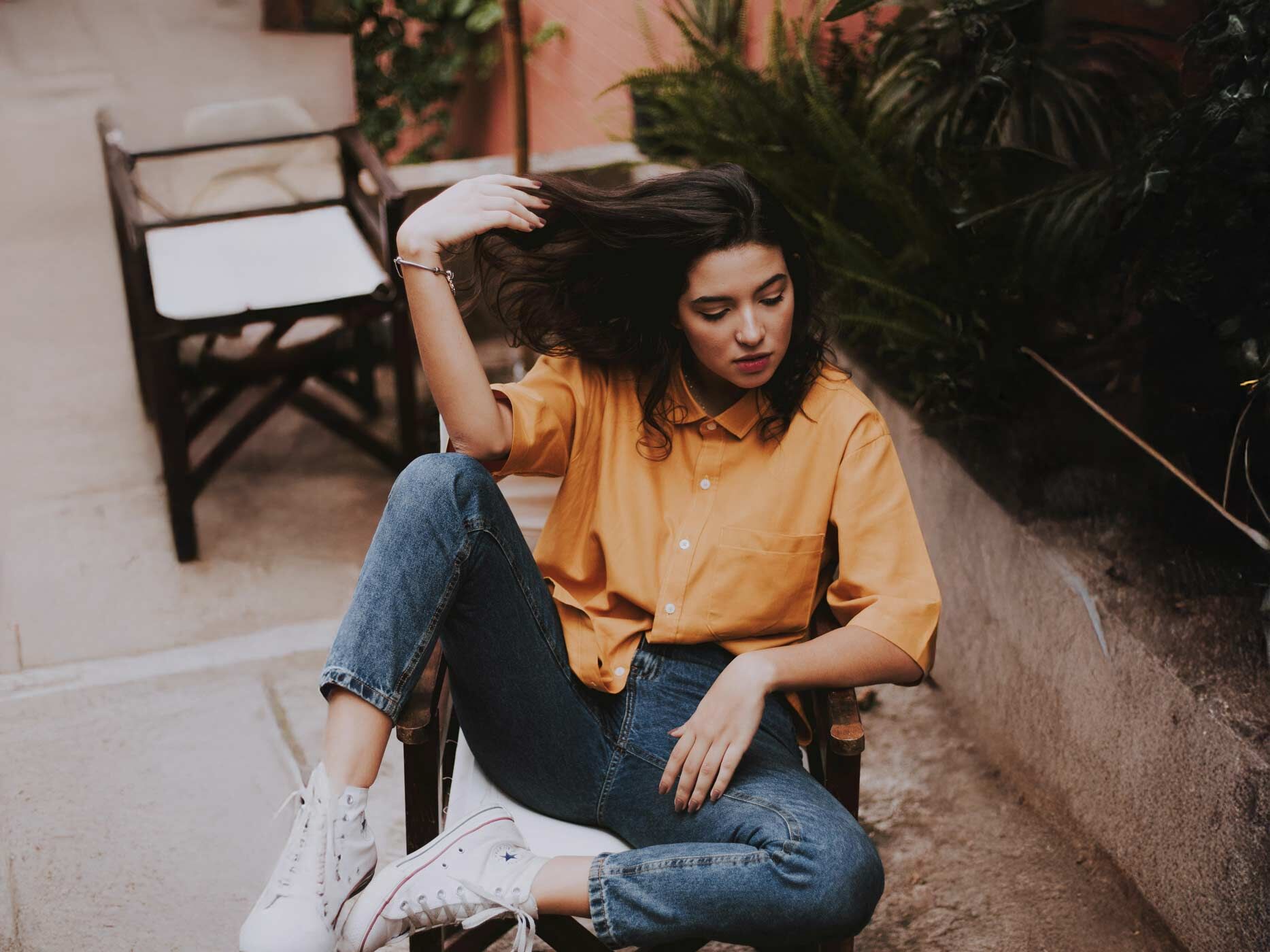Eine Frau sitzt auf einem Stuhl und trägt weiße Sneaker, eine dunkelblaue Jeans und ein locker geschnittenes, hellorangenes Hemd mit einer durchgehenden Knopfleiste.