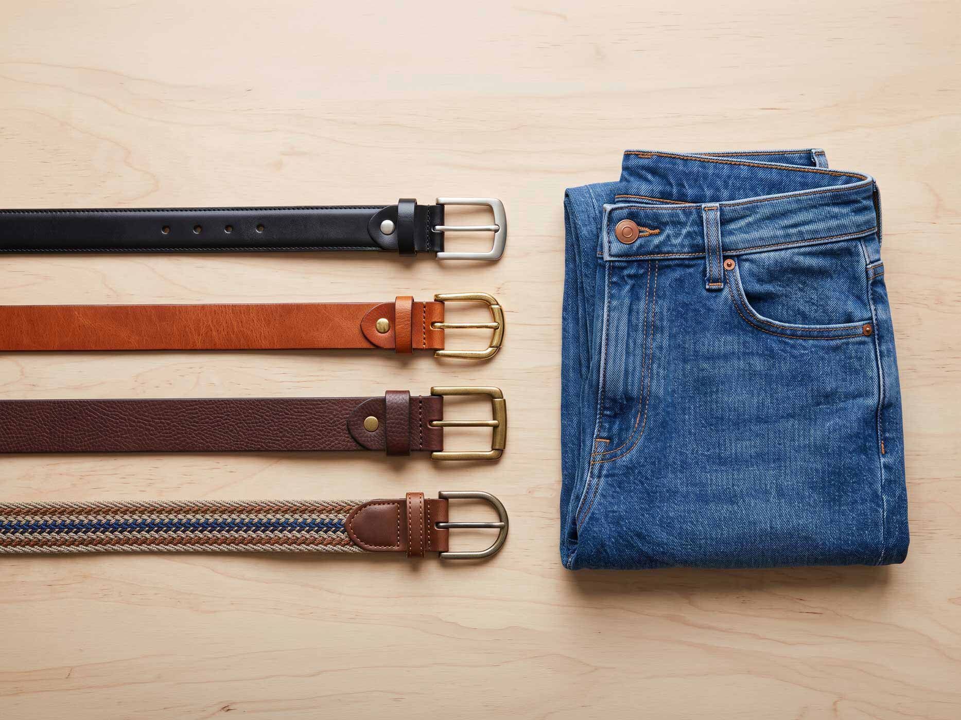 Eine gefaltete mittelblaue Jeans liegt neben vier verschiedenen Gürteln in Schwarz, Cognac, Dunkelbraun und mehrfarbiger Flechtoptik.