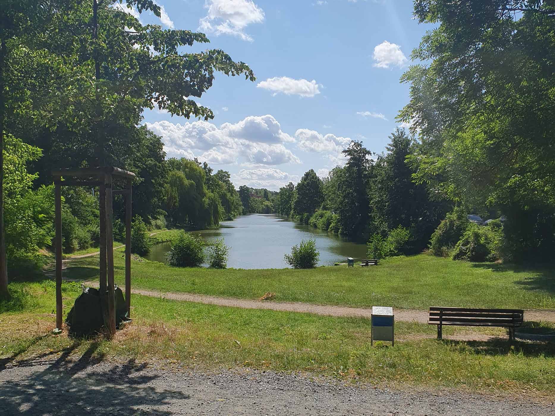 Ein gro&szlig;es Gew&auml;sser mit einem Spazierweg, einer Holzbank und vielen B&auml;umen drumherum.