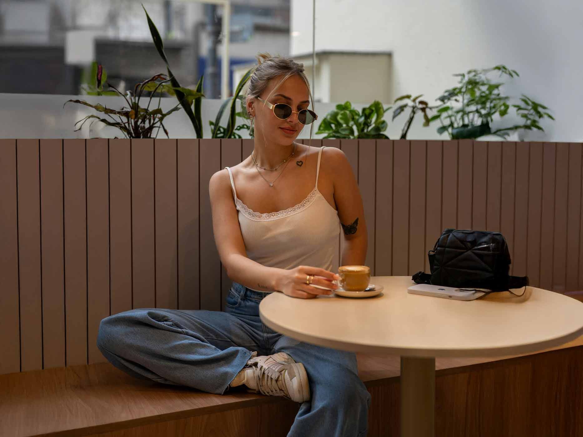 Eine Frau sitzt auf einer Bank an einem kleinen Tisch, sie trägt eine blaue Jeans, ein weißes Top mit einem Spitzenrand, goldenen Schmuck und eine Sonnenbrille.