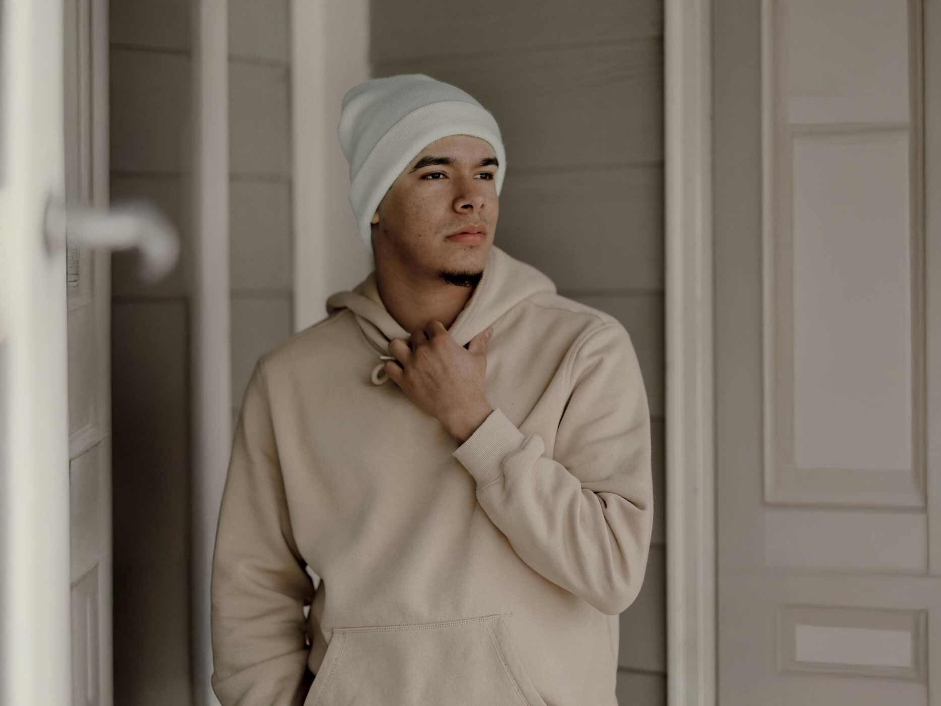 Ein Mann tr&auml;gt einen beigen Hoodie und eine wei&szlig;e M&uuml;tze, er guckt in die Ferne.