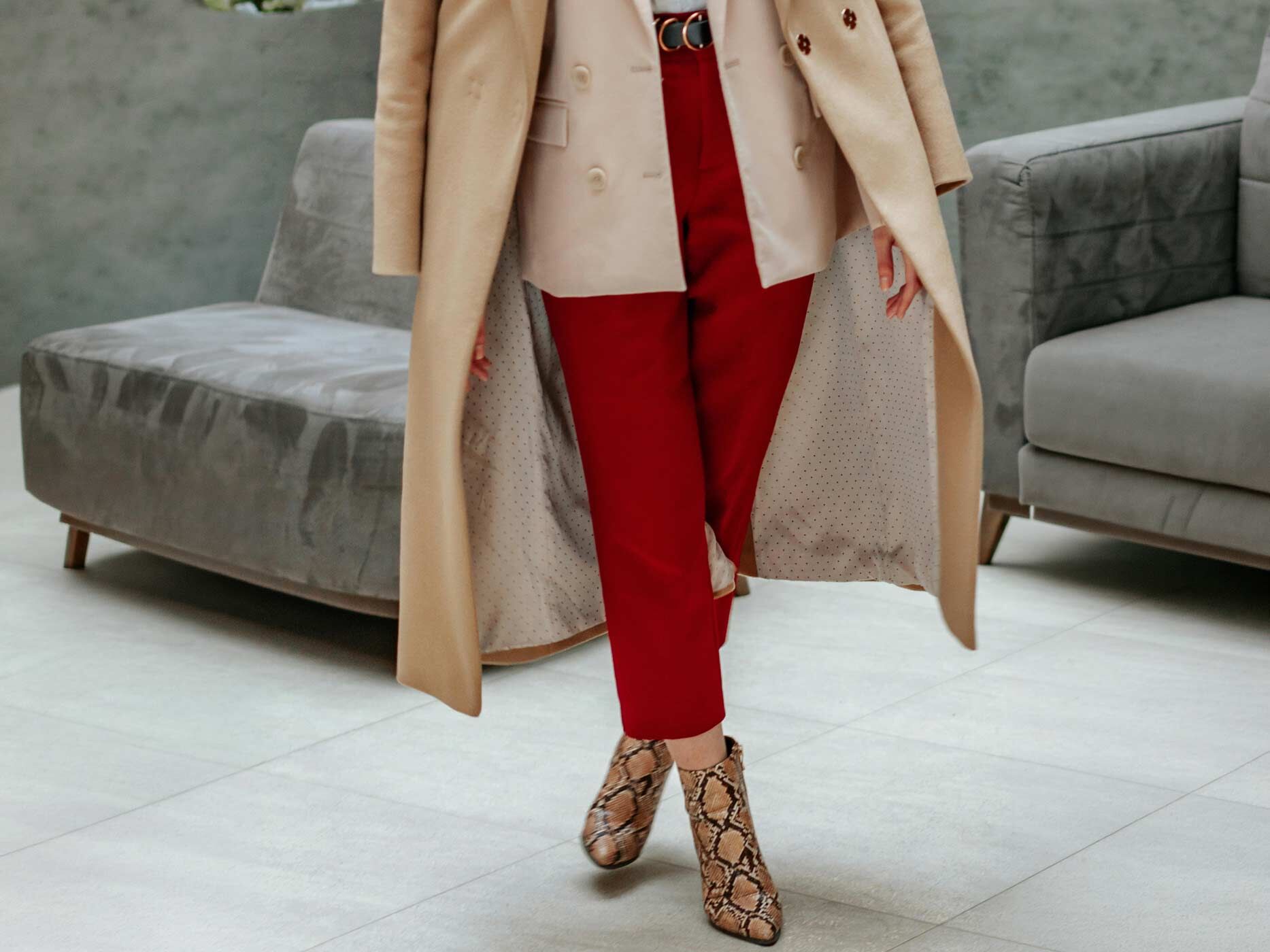 Eine Frau tr&auml;gt eine auff&auml;llige rote Hose, kombiniert mit einem hellen Oberteil und einem langen, beigen Mantel, dazu tr&auml;gt sie elegante Stiefeletten mit Schlangenoptik.