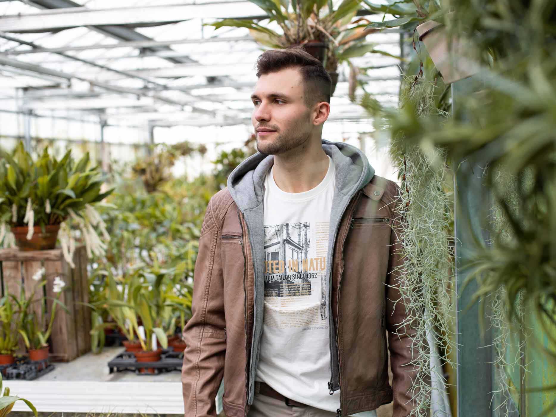 Ein Mann steht in einem Gewächshaus, er trägt ein weißes Oberteil, darüber eine graue Sweatjacke und darüber eine offene, hellbraune Lederjacke.