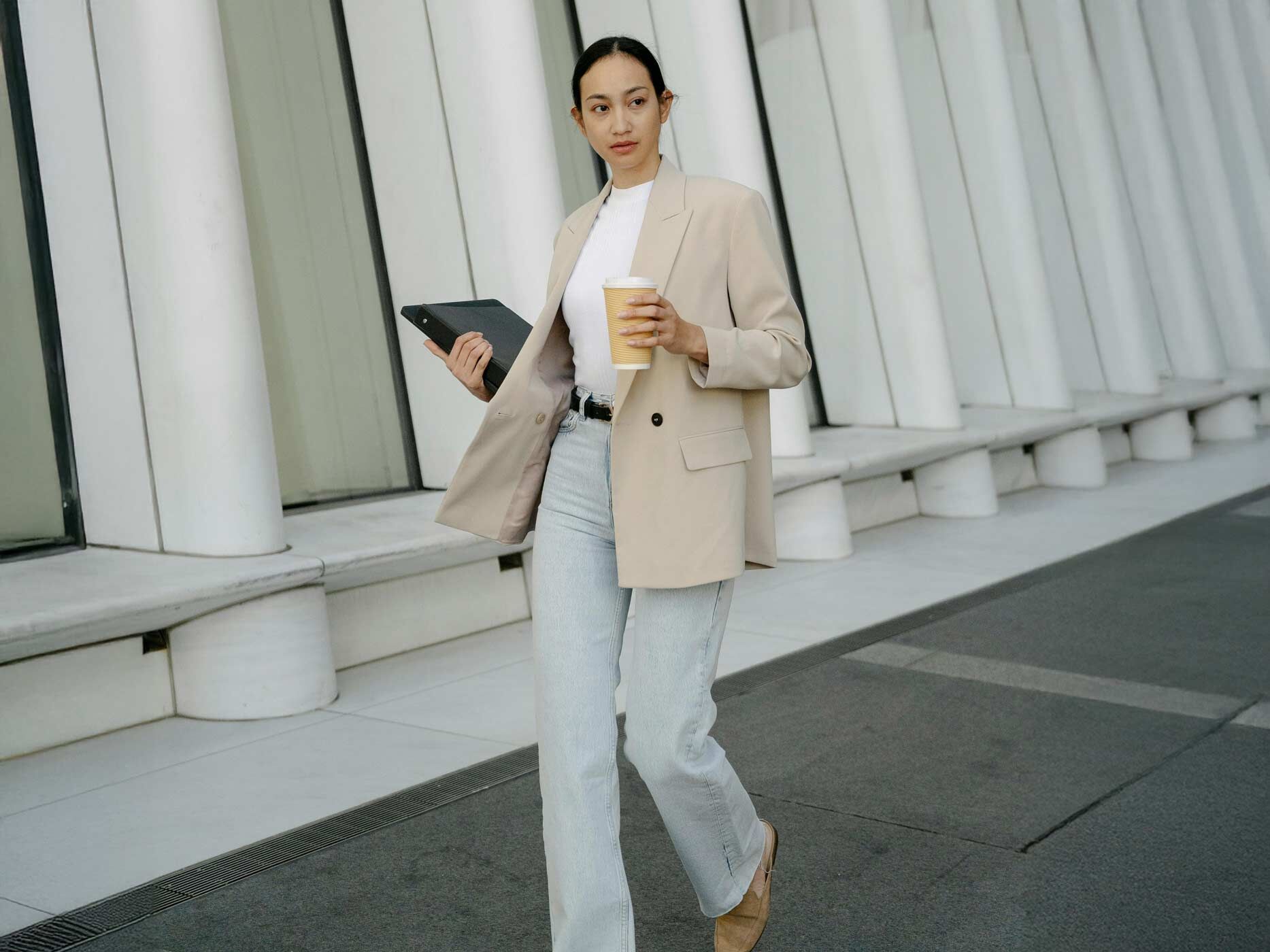 Eine Frau trägt eine hellblaue, locker geschnittene Jeans, dazu ein enges weißes Oberteil und einen Oversized beigen Blazer. Sie hält einen Kaffee und eine Aktentasche in der Hand und geht Treppen runter.