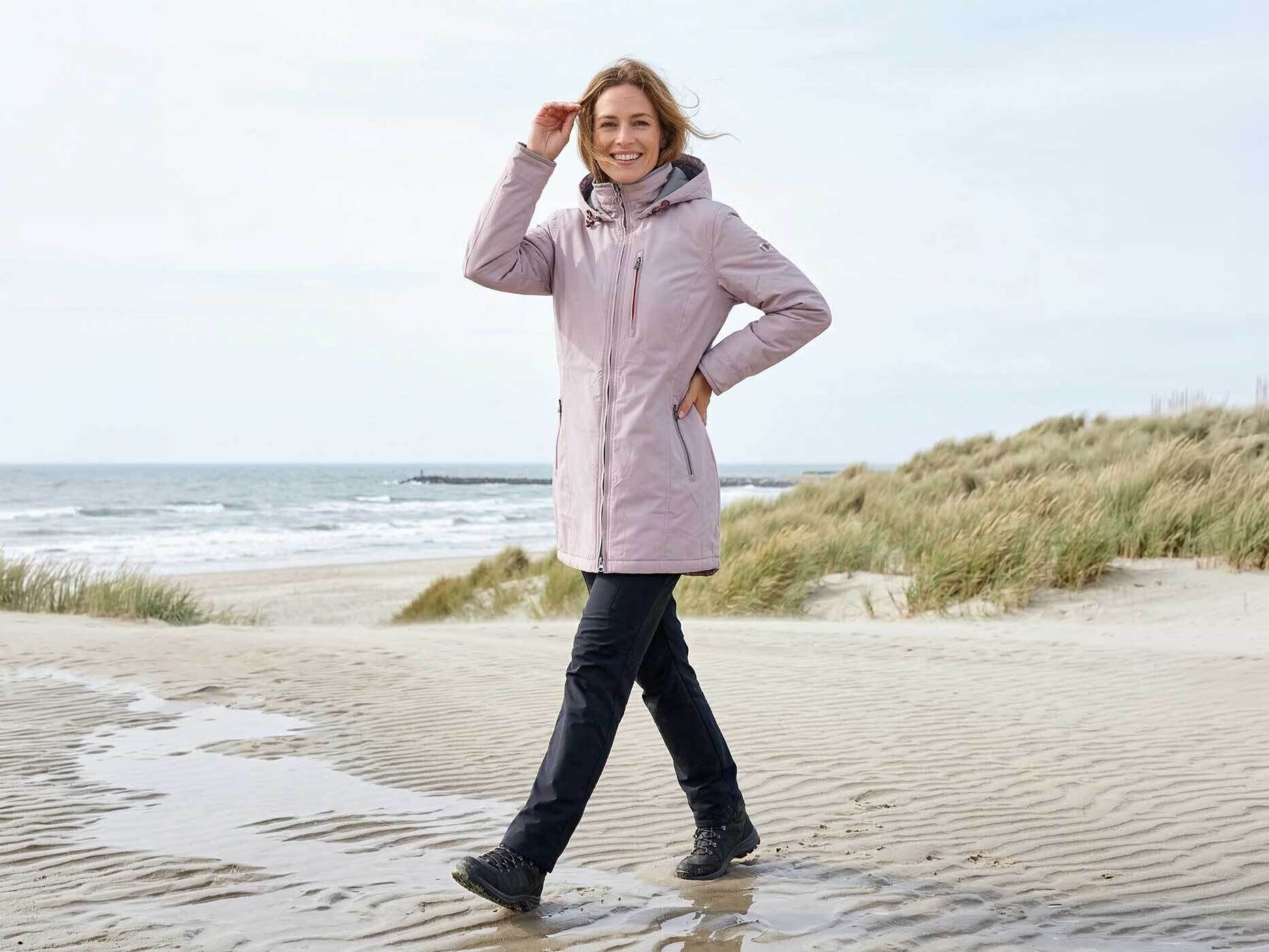 Eine Frau steht am Strand und tr&auml;gt eine Jacke in ashpink. Dazu tr&auml;gt sie eine blaue Hose.