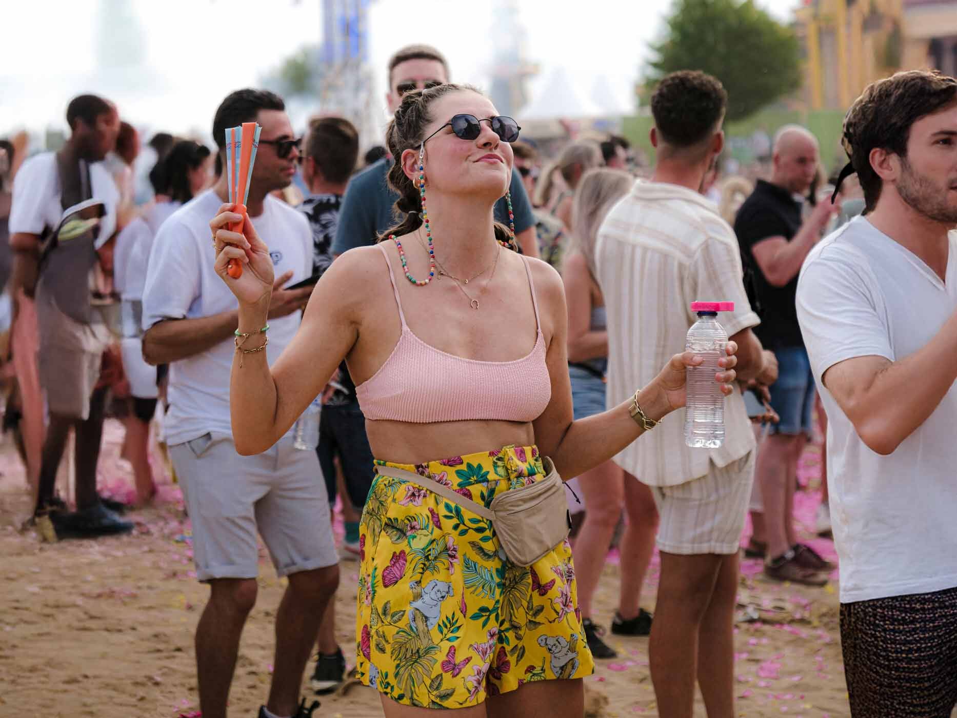 Eine Frau tanzt auf einem Festival, sie trägt eine gelbe Shorts mit Blumenprint, einen rosafarbenen BH, eine schwarze Sonnenbrille und hat dabei einen Fächer und eine Wasserflasche in der Hand.