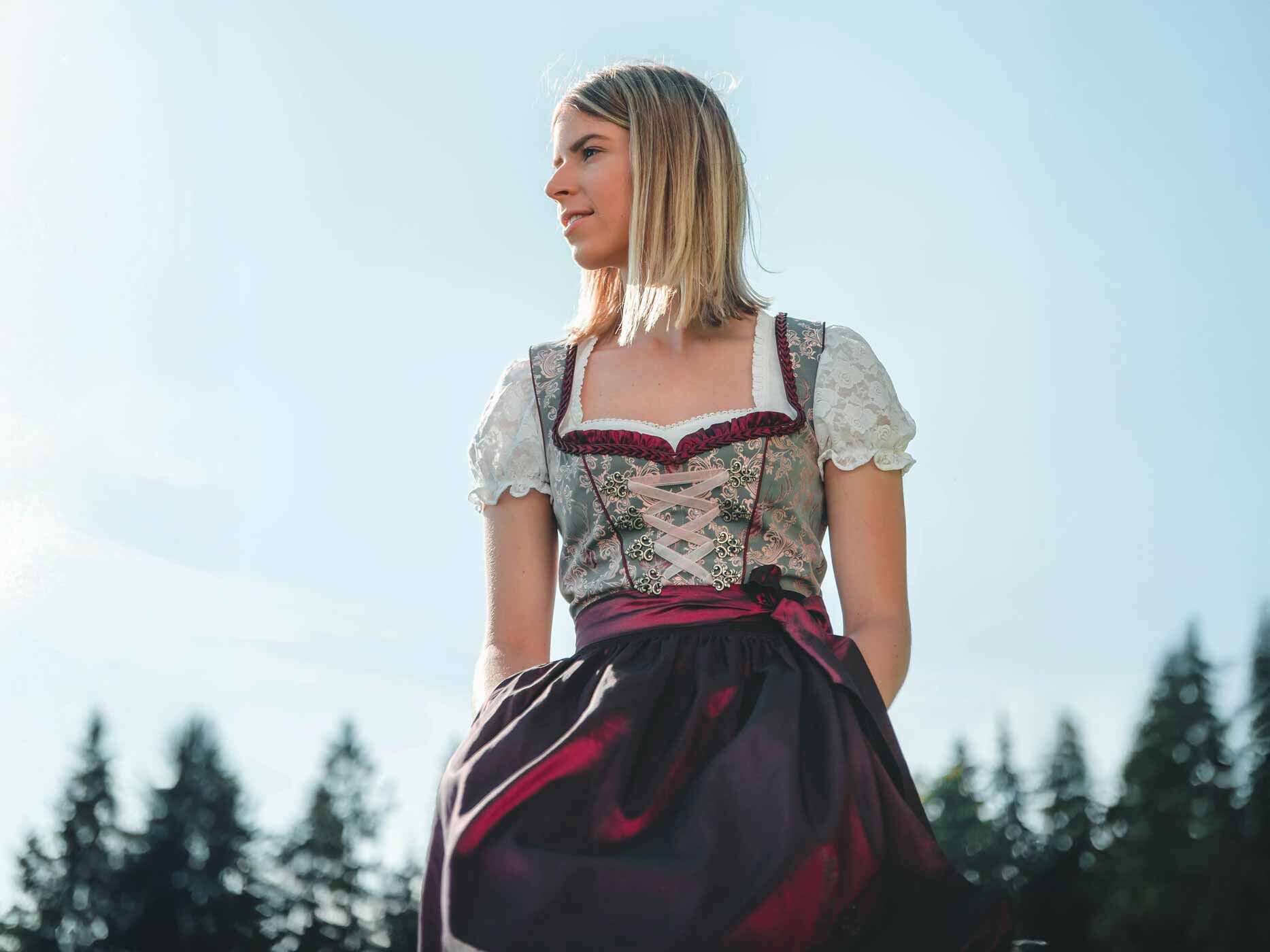 Eine Frau trägt eine weiße Bluse, ein grünes Dirndl-Oberteil und eine dunkelrote Schürze.