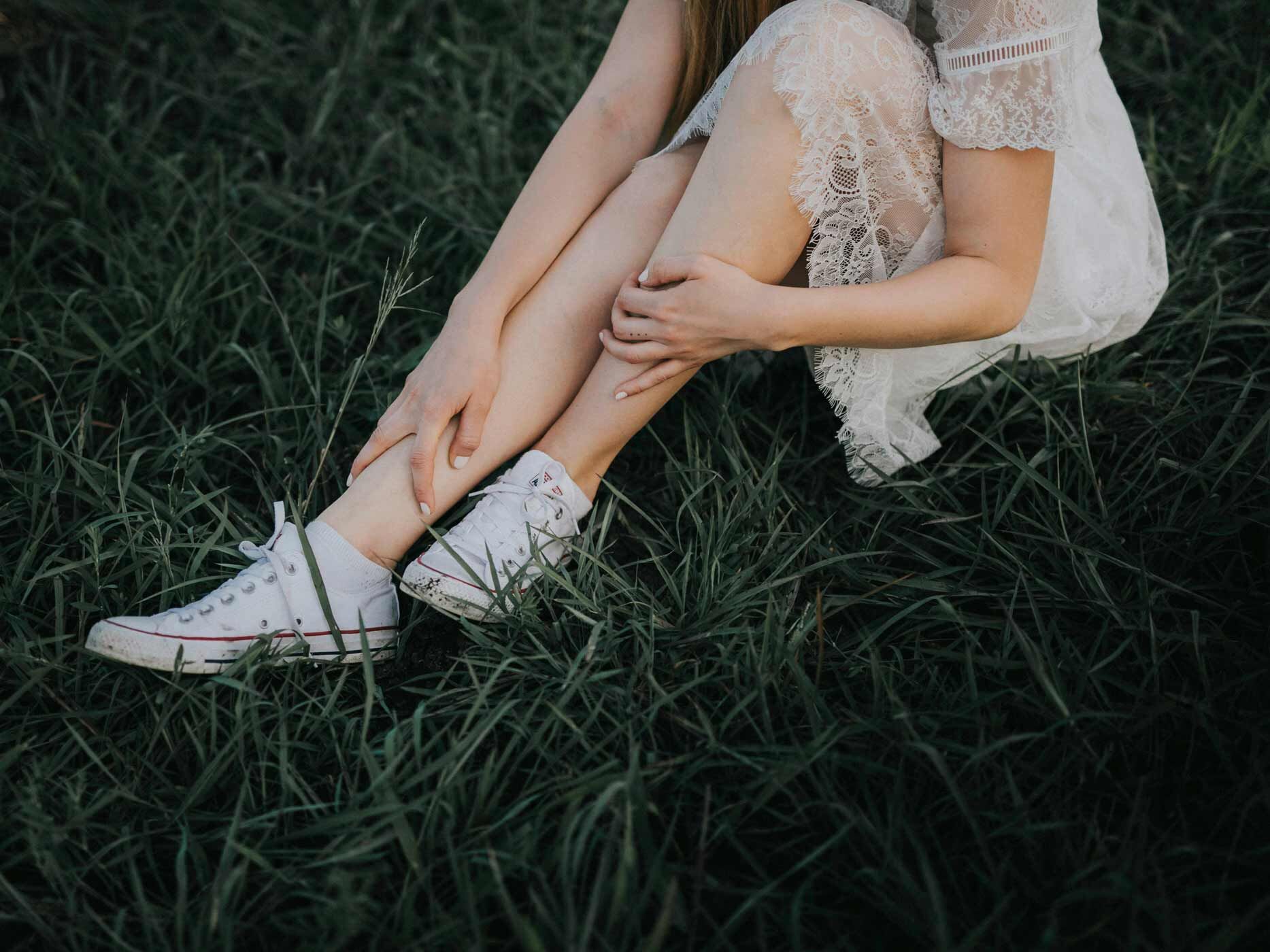 Eine Frau sitzt im Gras und trägt ein kurzes, weißes Sommerkleid und weiße Chucks.