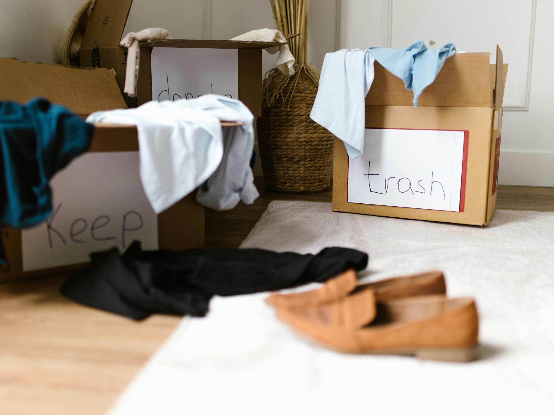 Drei Kartons mit den Aufschriften "Keep", "Trash" and "Donate" stehen auf dem Boden, drum herum sind Klamotten verteilt.