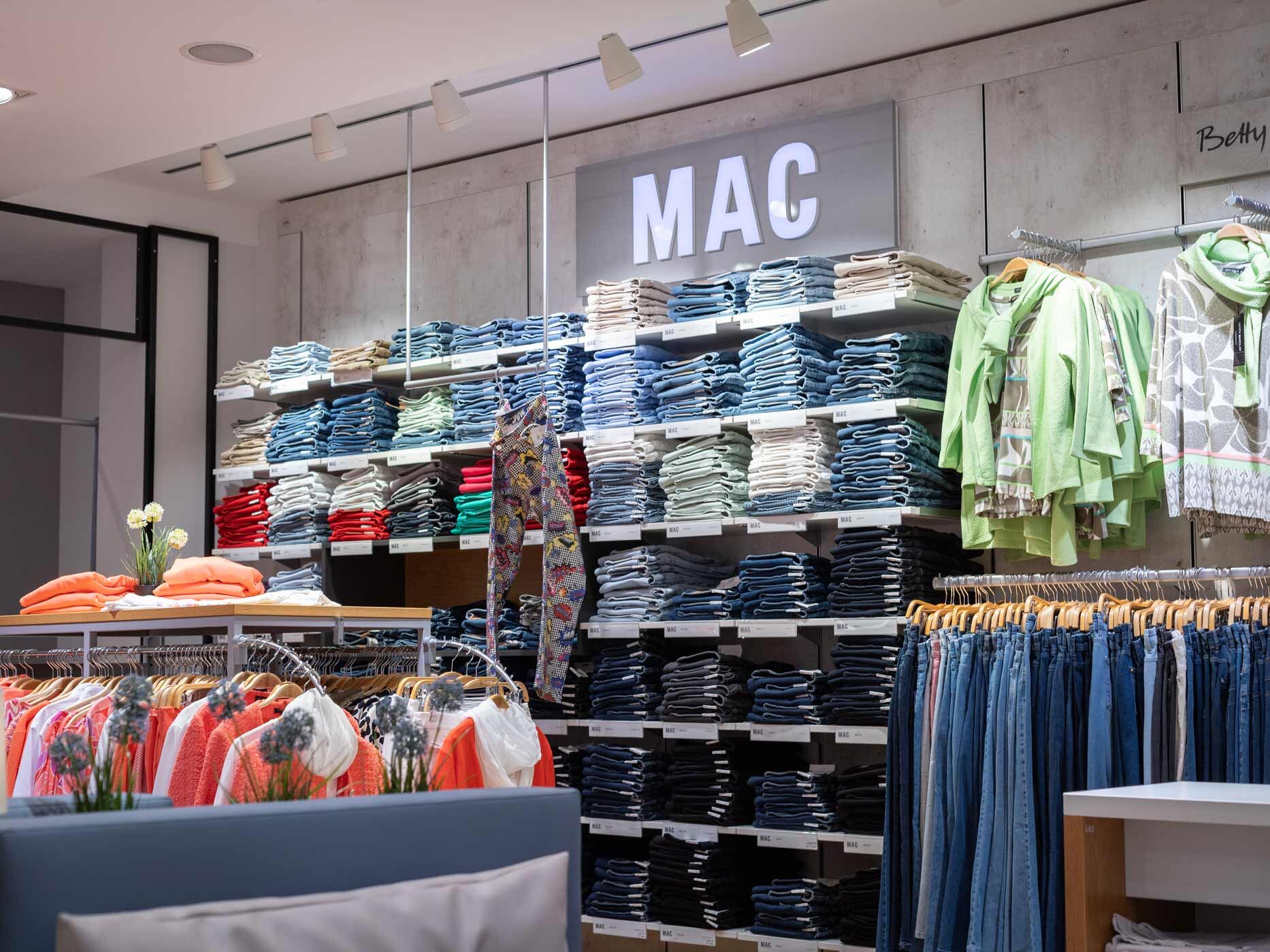 Große Jeansabteilung mit einer umfangreichen Auswahl an Damenjeans der Marke MAC. Die Regale sind mit unterschiedlich gewaschenen Modellen gefüllt, ergänzt durch farbige Oberteile und Accessoires.