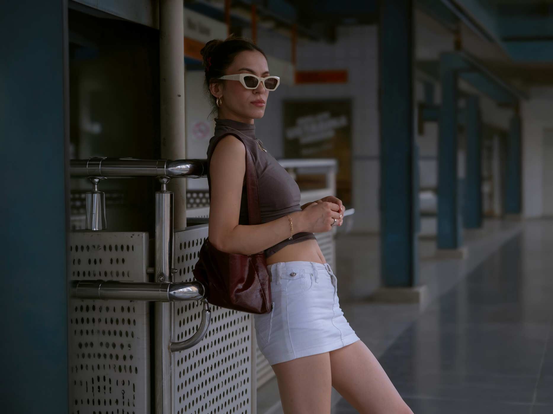Eine Frau lehnt an ein Gel&auml;nder, sie tr&auml;gt einen wei&szlig;en Minijeansrock, ein braunes Top, eine braune Handtasche, goldenen Schmuck und eine beige Sonnenbrille.