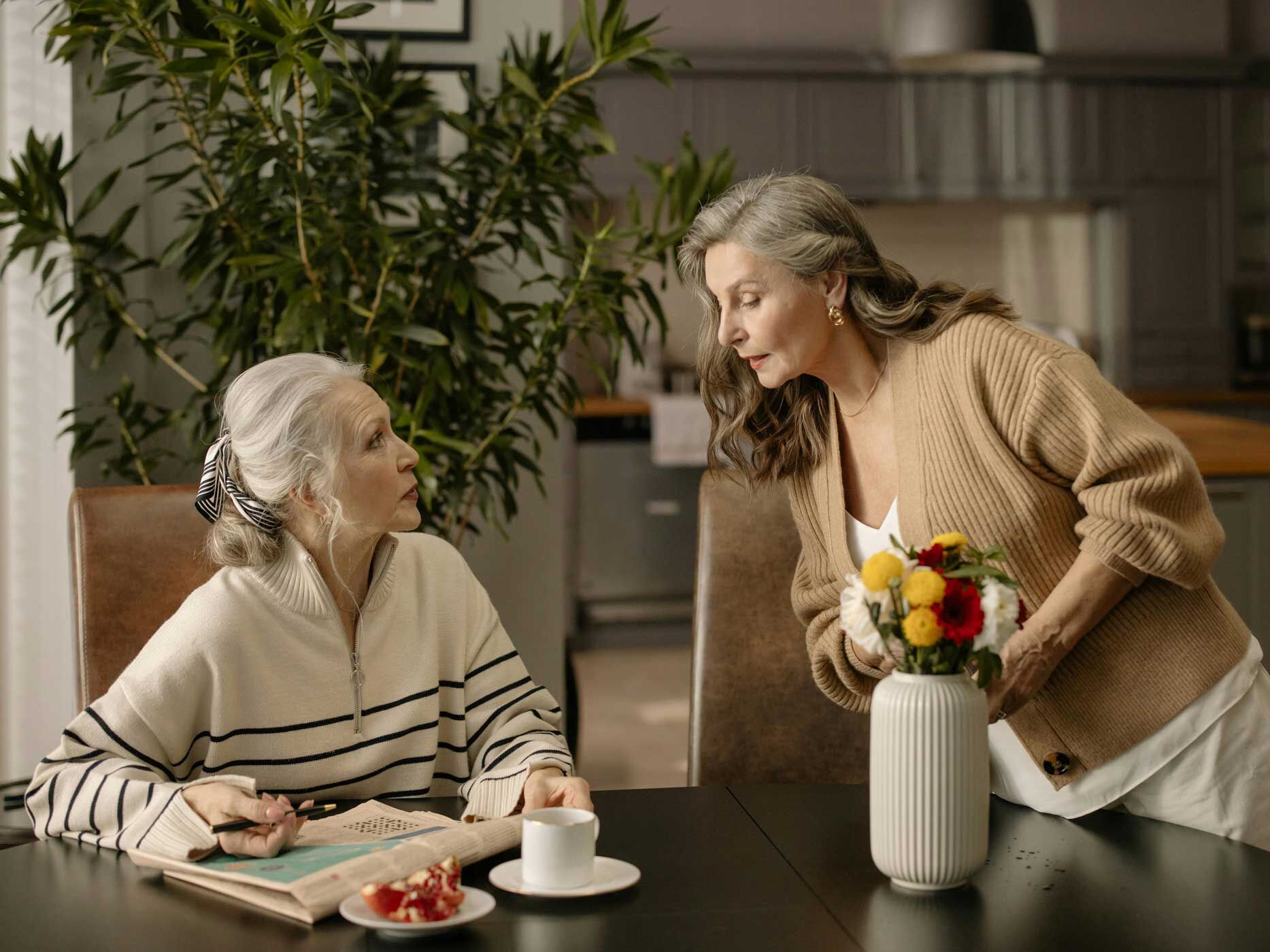 Zwei &auml;ltere Frauen mit grauen Haaren tragen ruhige, hochwertige Kleidung in Naturt&ouml;nen. Eine sitzt in einem cremefarbenen Pullover mit dunklen Streifen, die andere tr&auml;gt einen beigefarbenen Cardigan &uuml;ber einem hellen Oberteil, beide Outfits wirken weich, klassisch und zeitlos.
