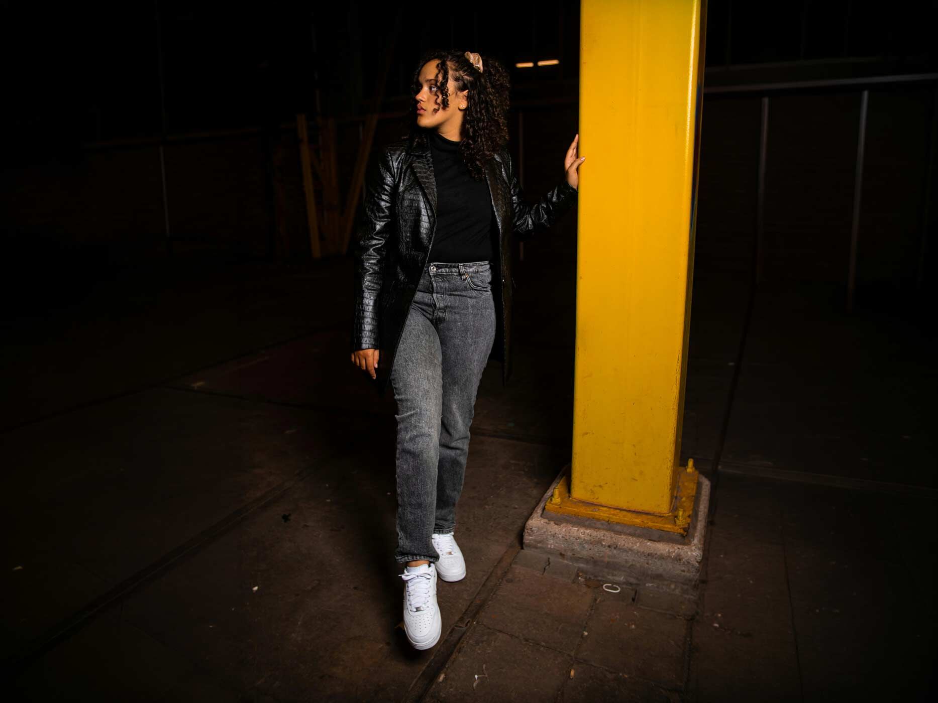 Eine Frau steht in einem Parkhaus, sie tr&auml;gt eine graue Jeans, ein schwarzes Oberteil und dar&uuml;ber eine schwarze Lederjacke und wei&szlig;e Sneaker.