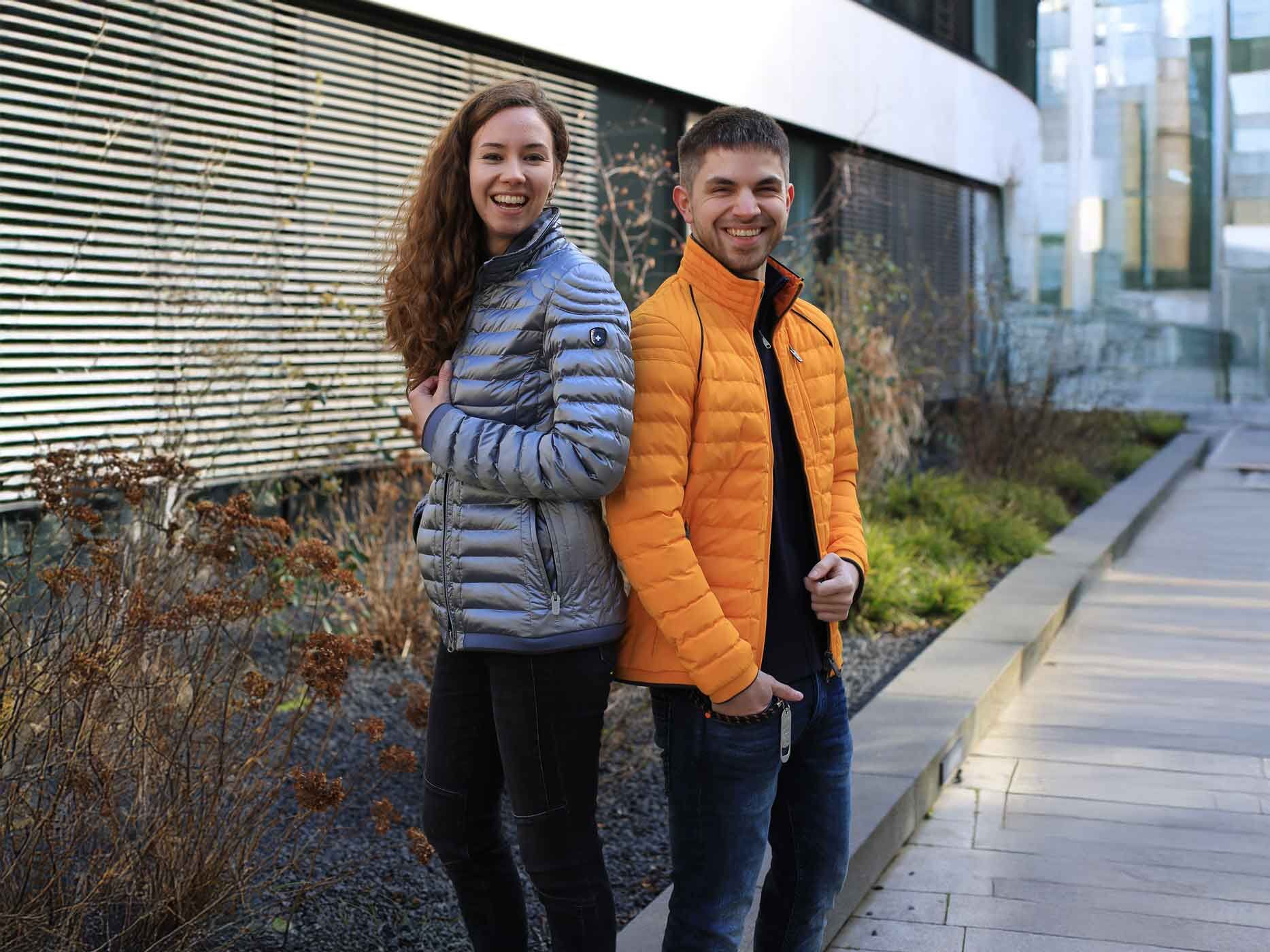 Ein Mann und eine Frau stehen Rücken an Rücken und lachen in die Kamera, sie trägt eine graue Wellensteyn Jacke und er eine Orangene.