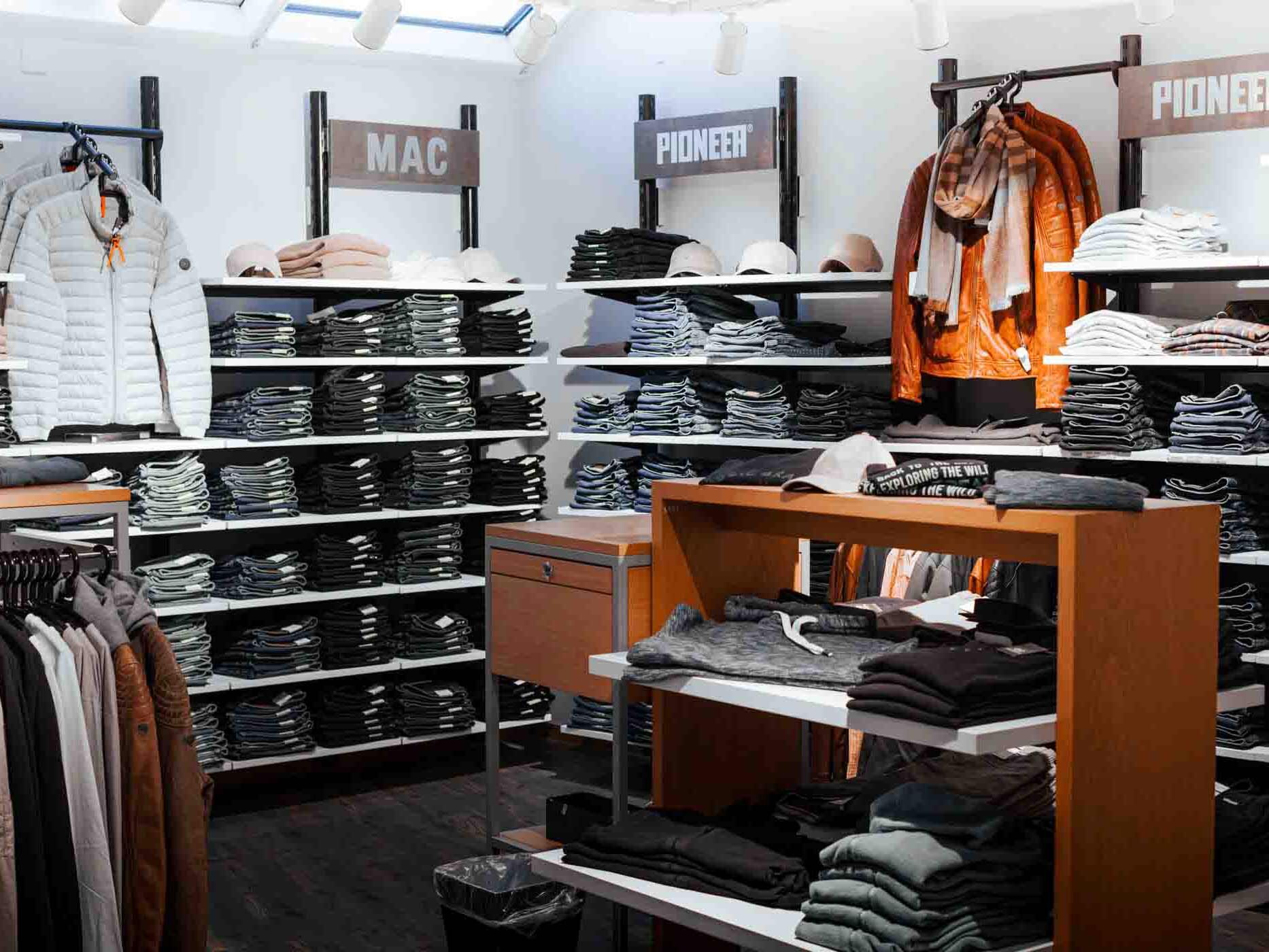 Herrenabteilung mit Jeans und Freizeitmode der Marken MAC und Pioneer. Dunkle Jeansstapel, Jacken und Strickwaren sind &uuml;bersichtlich auf Regalen und Verkaufstischen pr&auml;sentiert.