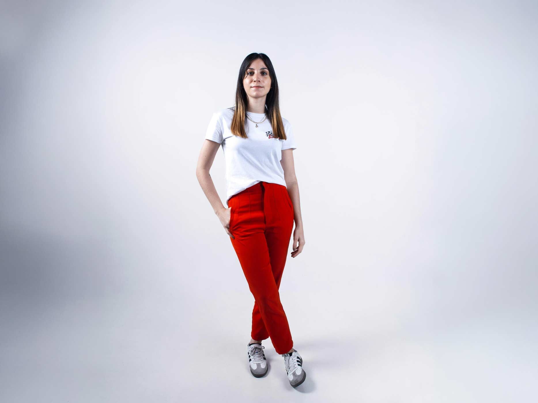 Eine Frau tr&auml;gt eine rote Stoffhose, dazu ein wei&szlig;es T-Shirt mit Print, das in die Hose gesteckt ist, wei&szlig;-graue Sneaker und eine Kette.