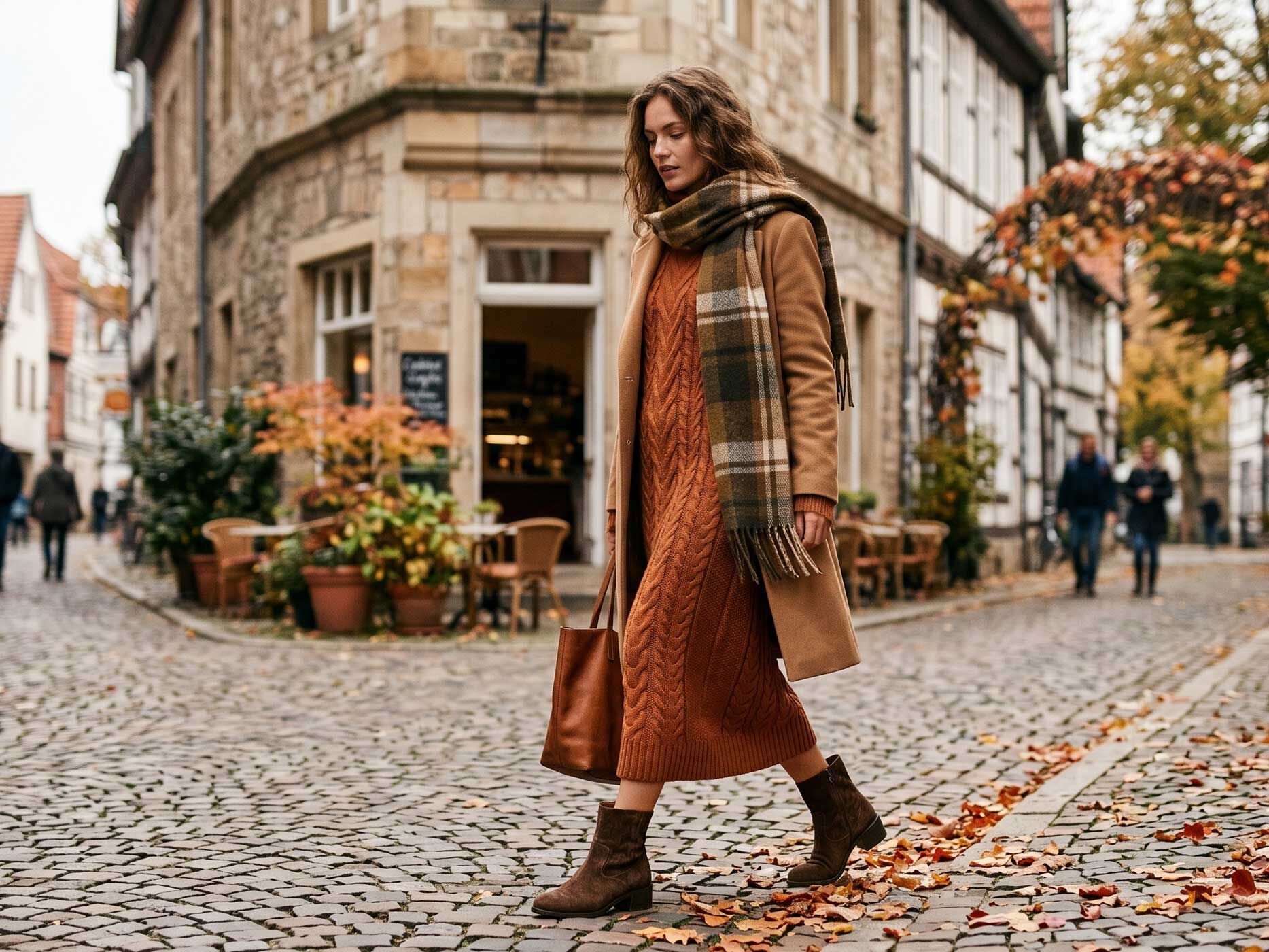 Eine Frau kombiniert ein rostbraunes Strickkleid mit camelfarbenem Mantel, kariertem Schal und braunen Stiefeletten.