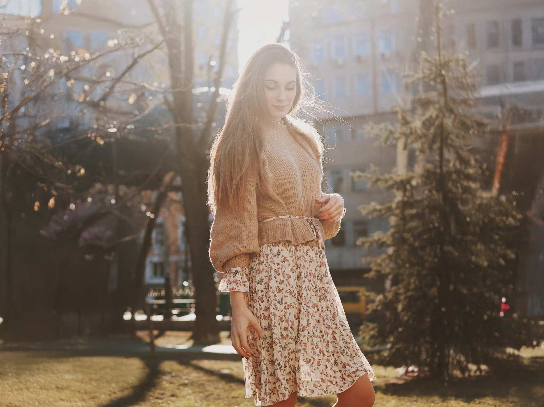 Eine Frau trägt ein weißes Sommerkleid mit Blumenmuster und langen Ärmeln, dazu trägt sie einen braunen Pullover.