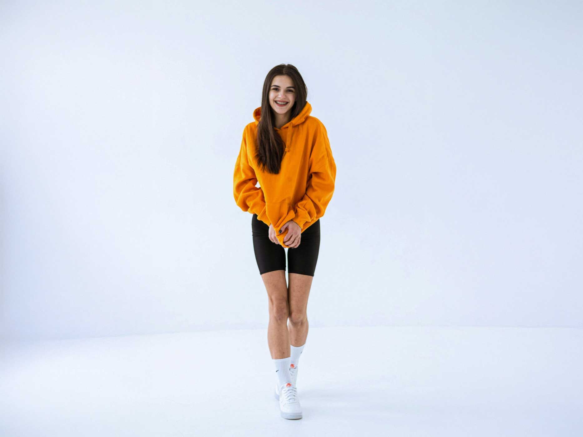 Eine junge Frau tr&auml;gt eine schwarze Radlerhose, dar&uuml;ber einen locker geschnittenen orangene Hoodie, wei&szlig;e Tennissocken und wei&szlig;e Sneaker.