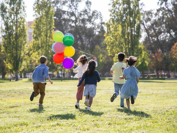 Kinder Laufen über eine Wiese und halten dabei Luftballons in ihren Händen. 