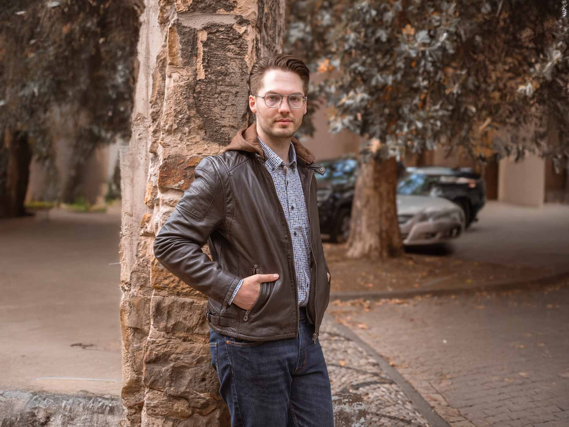 Ein Mann lehnt an einen Baum, er trägt eine dunkelblaue Jeans, ein gepunktetes Hemd und eine offene braune Lederjacke darüber.